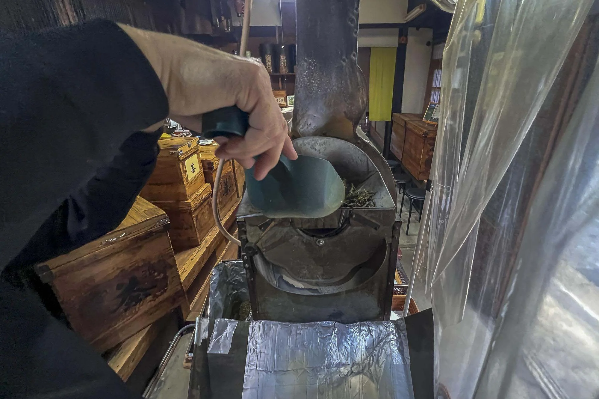A person holding a scoop above a large metal container, possibly in a workshop or craft space, with wooden furniture and other equipment in the background.