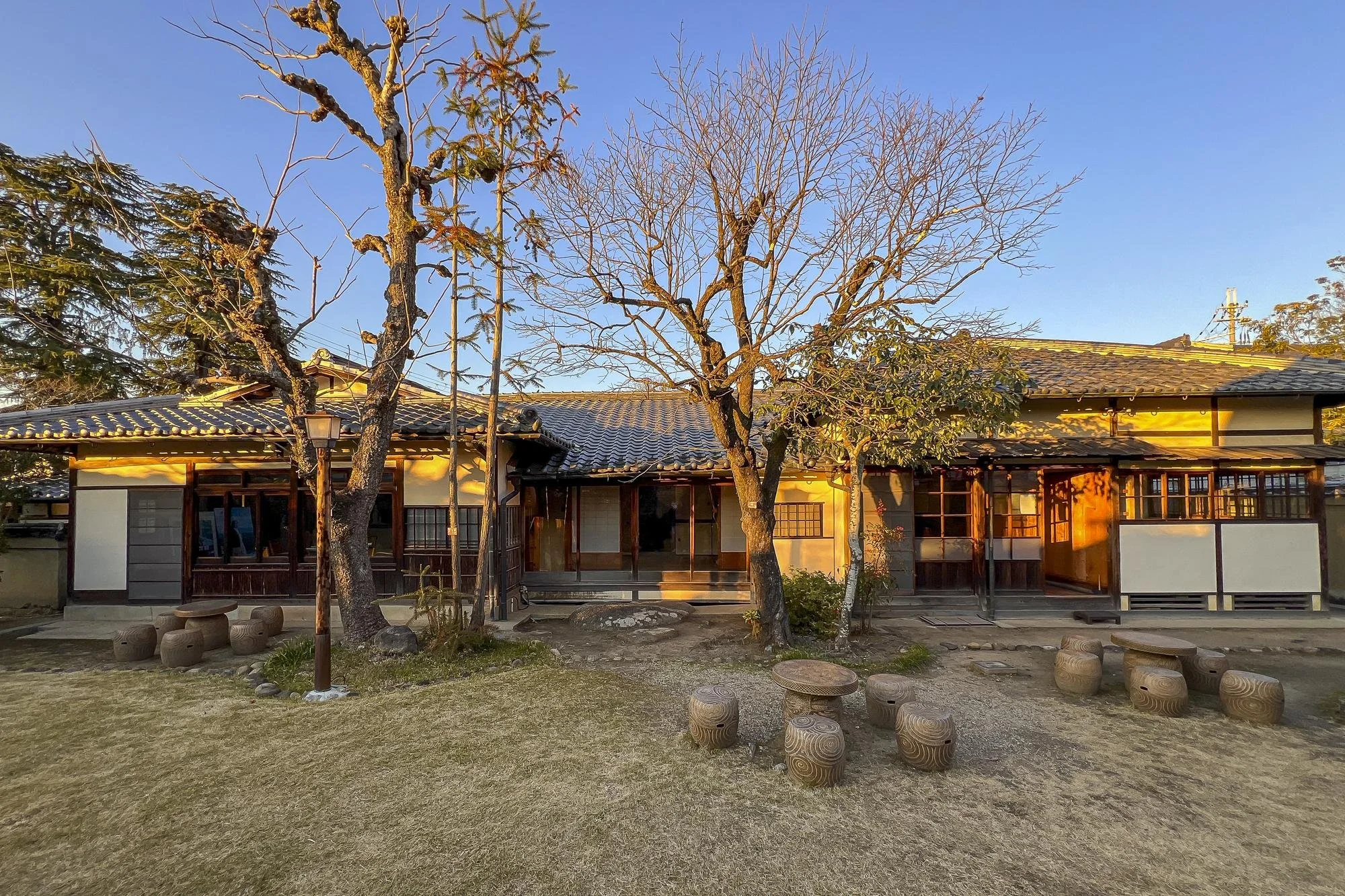 Traditional Japanese house with wooden exterior, surrounded by trees and outdoor seating area with wooden stools and a table, illuminated by warm sunlight during sunset.