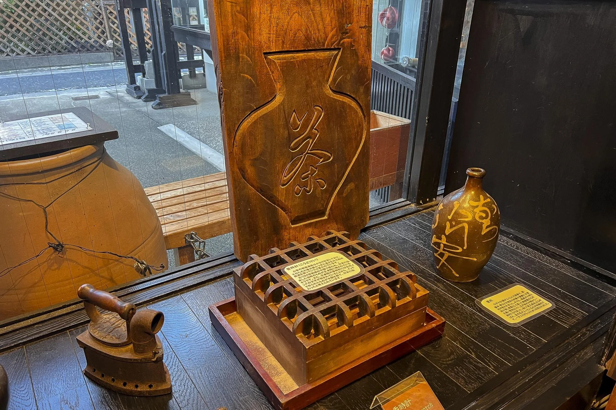 Display of traditional Japanese objects including a wooden sign with calligraphy, an antique iron, a decorative vase with Japanese characters, and informational signs, all inside a store with a window showing the street outside.