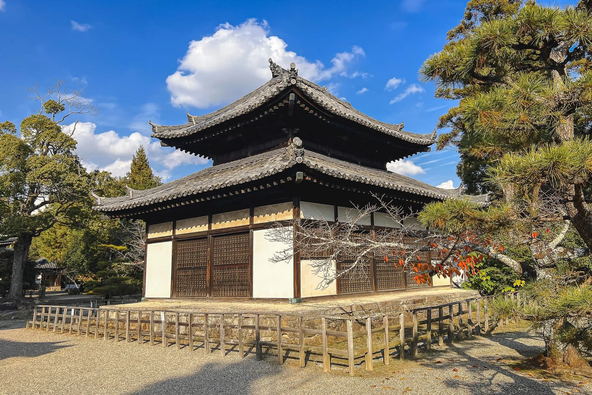 Traditional Japanese building with wooden sliding doors, surrounded by trees and a gravel pathway, under a blue sky with white clouds.