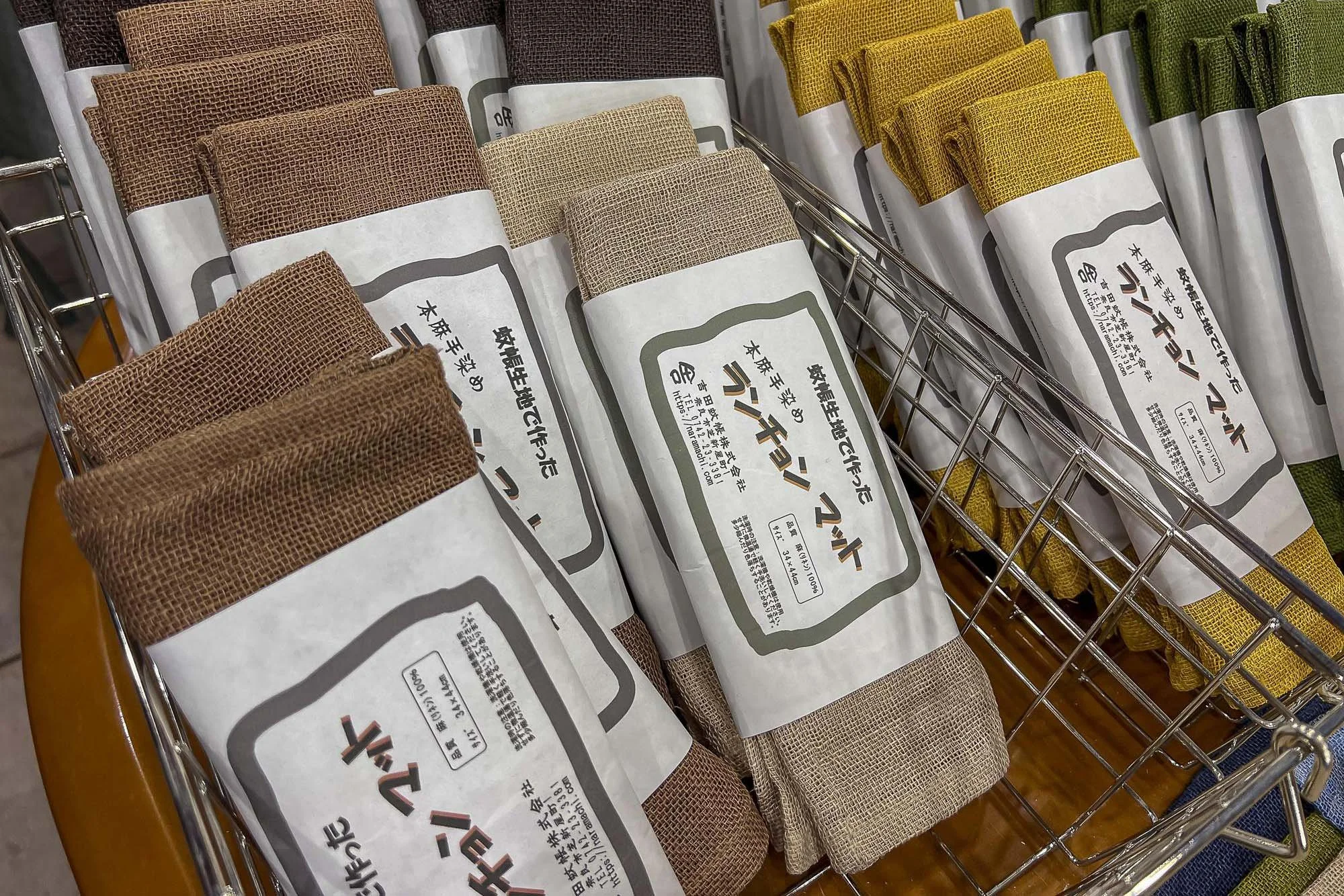 rolls of woven fabric in various earth-tone colors, labeled in Japanese, placed in a metal shopping basket.