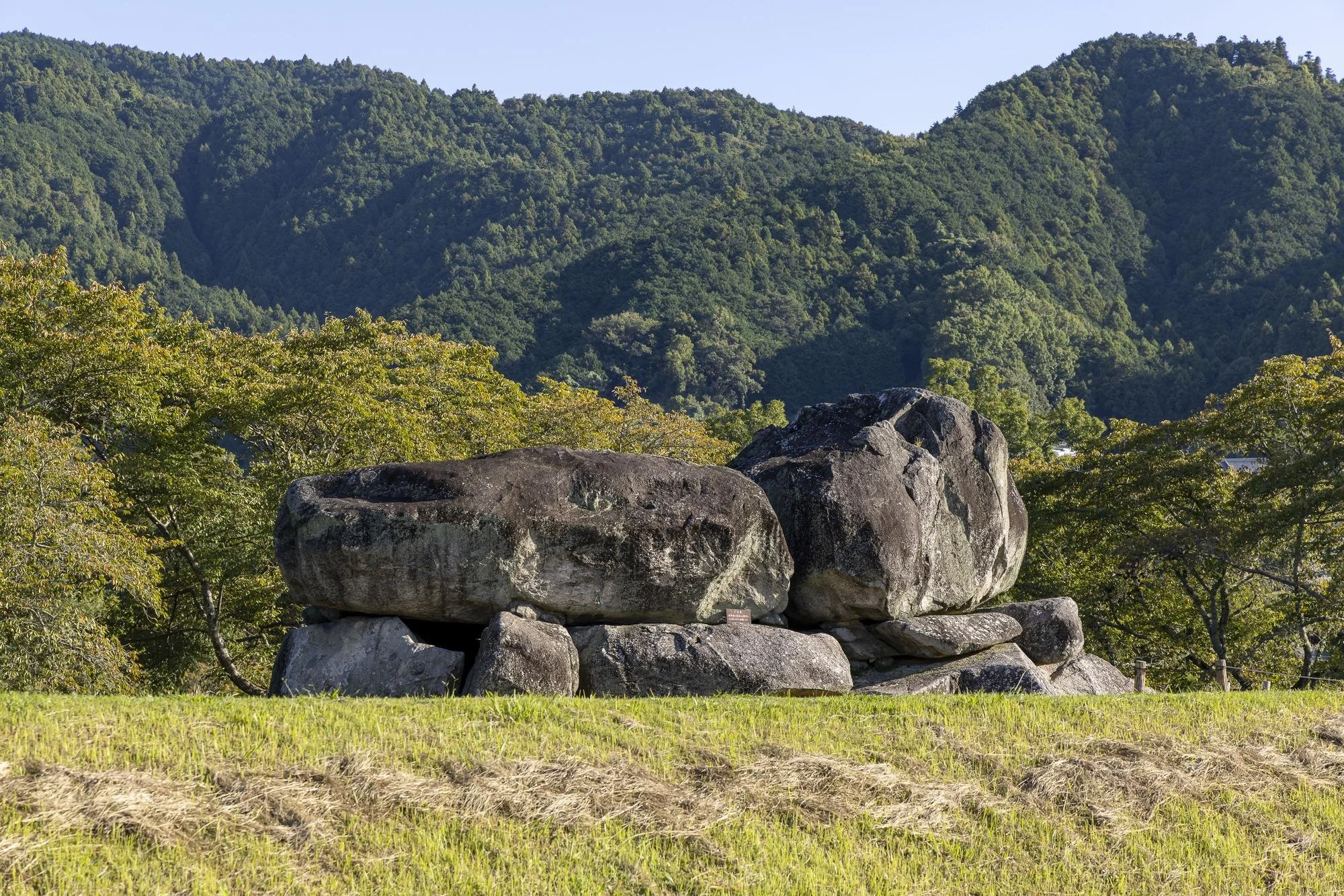 Ishibutai Mounded Tomb: Large ancient stone formation in a grassy clearing, with green forested mountains in the background.