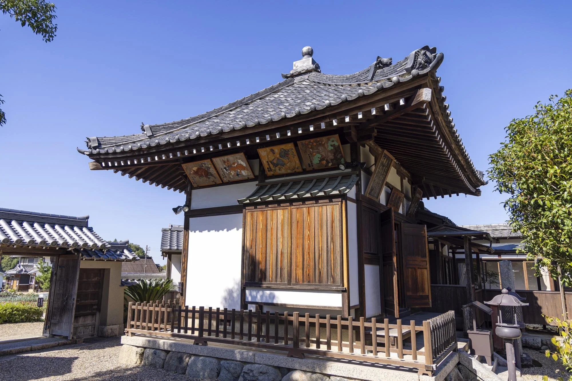 Traditional Japanese wooden building with tiled roof, sliding wooden doors, and decorative paintings on the eaves, surrounded by a small wooden fence and greenery, under a clear blue sky.