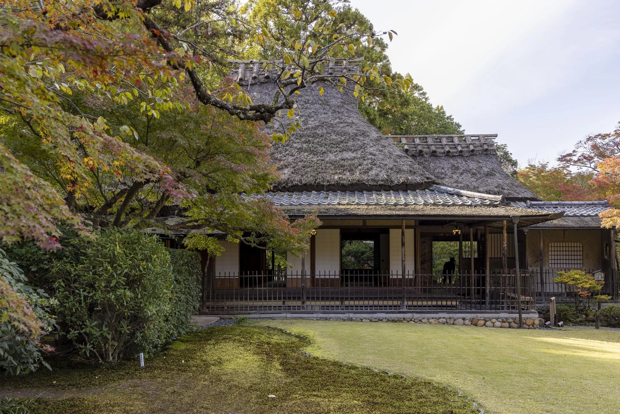 Yoshikien Garden: Traditional Japanese thatched-roof house surrounded by trees with autumn leaves and a maintained grass lawn.