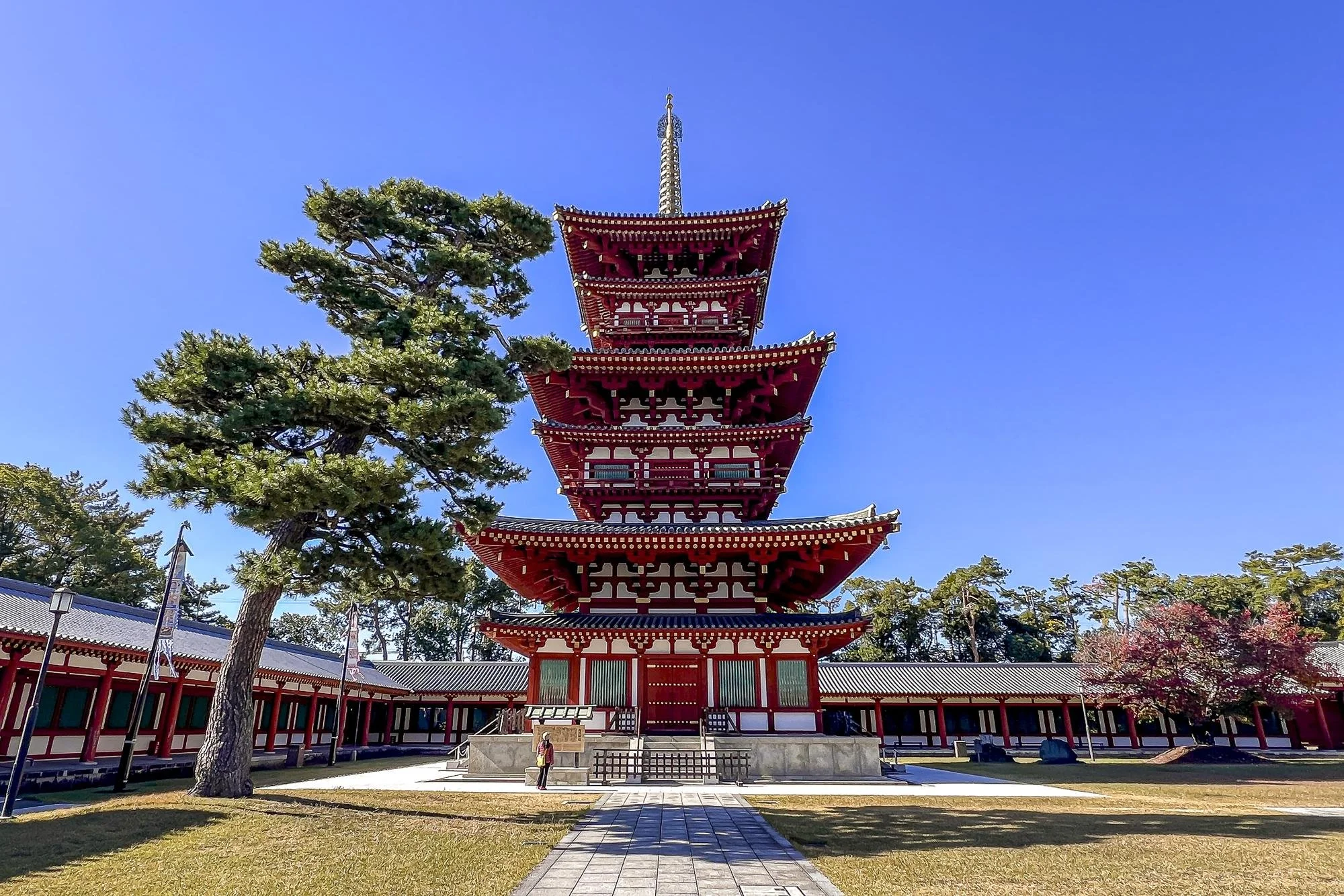 A traditional Japanese five-story pagoda with red and white colors, surrounded by trees, clear blue sky, and a person standing in front.