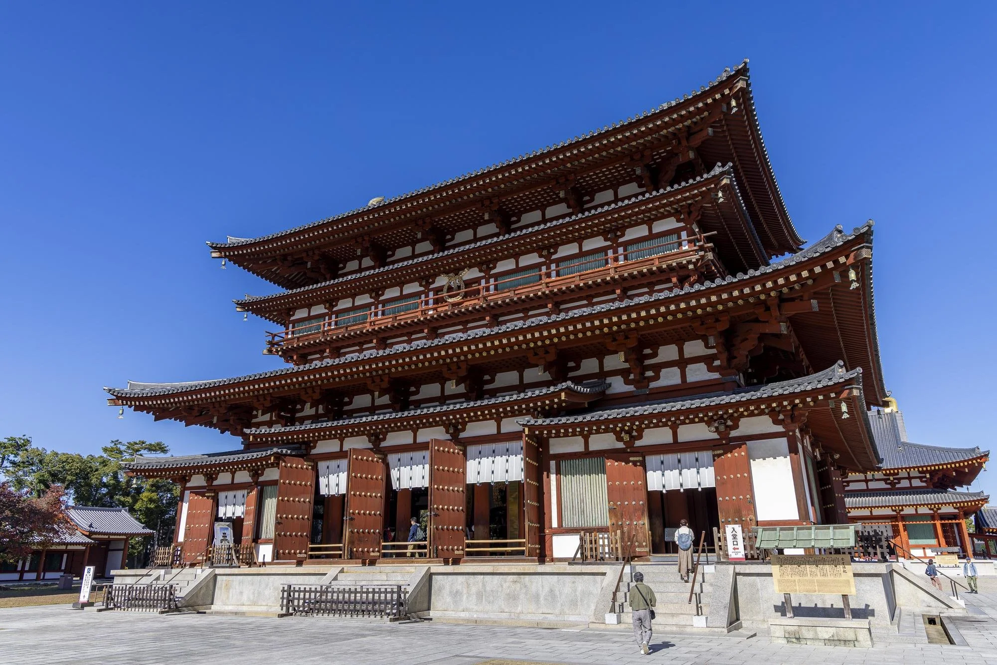 Traditional Japanese temple with multi-tiered roof structure, wooden beams, and ornate design, set against a clear blue sky.