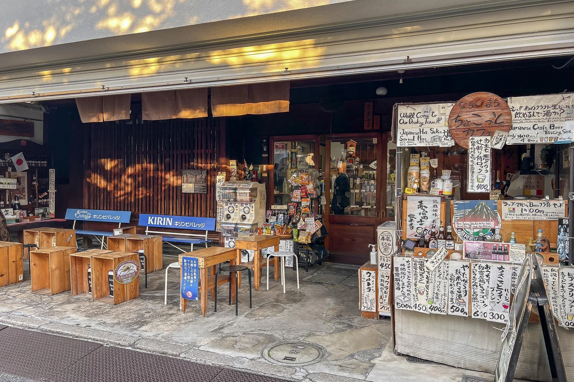Japanese storefront with outdoor seating, advertising signs, and menu boards displaying prices in Japanese yen, and various beverages and snacks on display.