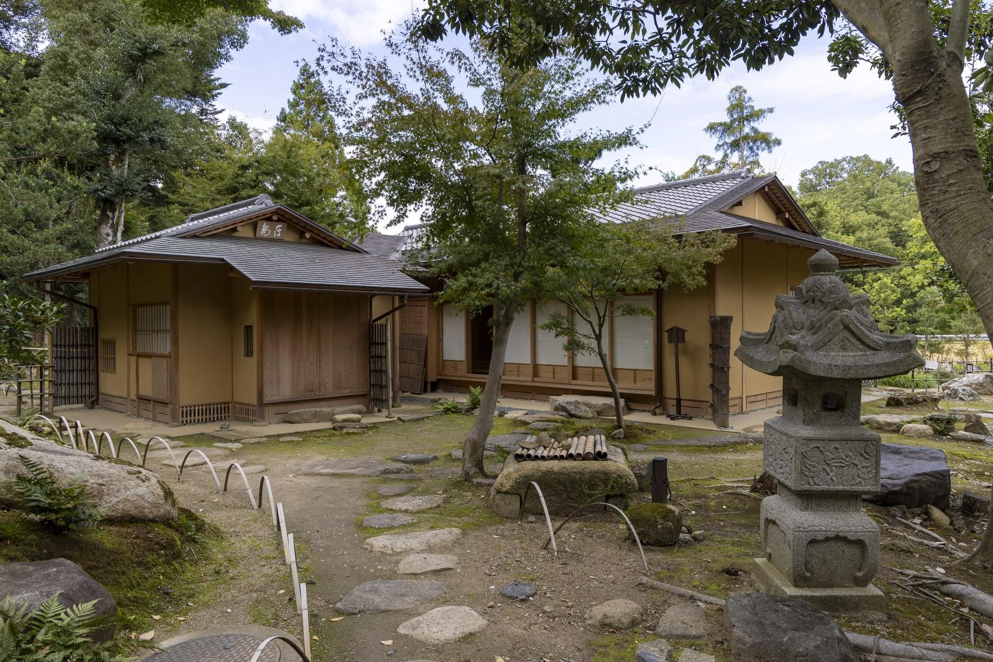 Traditional Japanese tea house with a stone lantern and a garden path surrounded by trees and rocks.