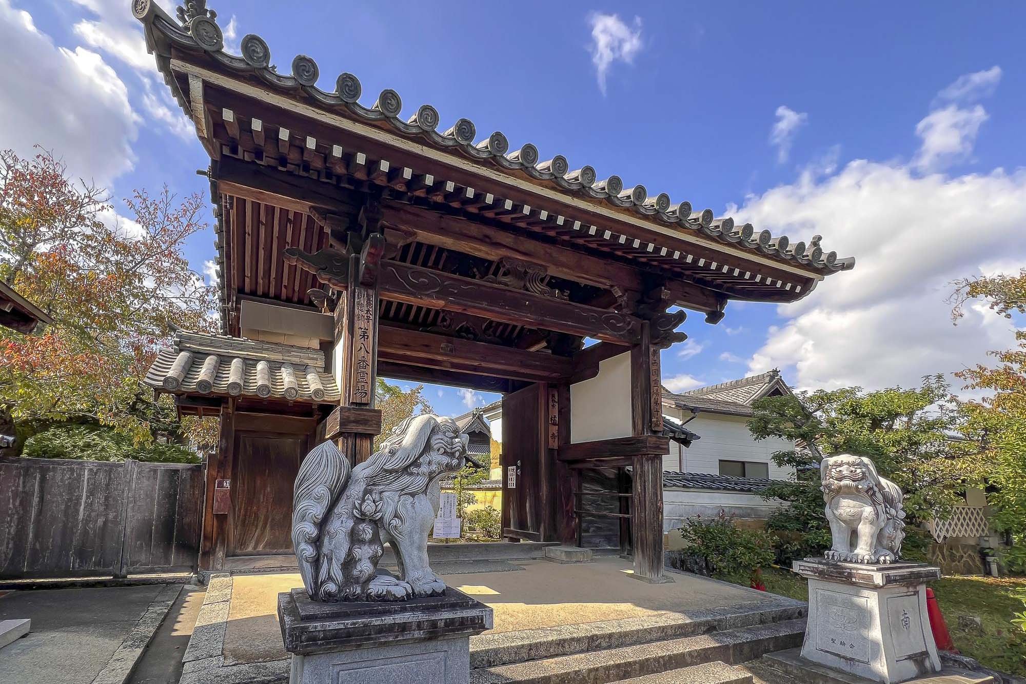 Traditional Japanese temple gate with two stone lion statues at the entrance, under a blue sky with some clouds.