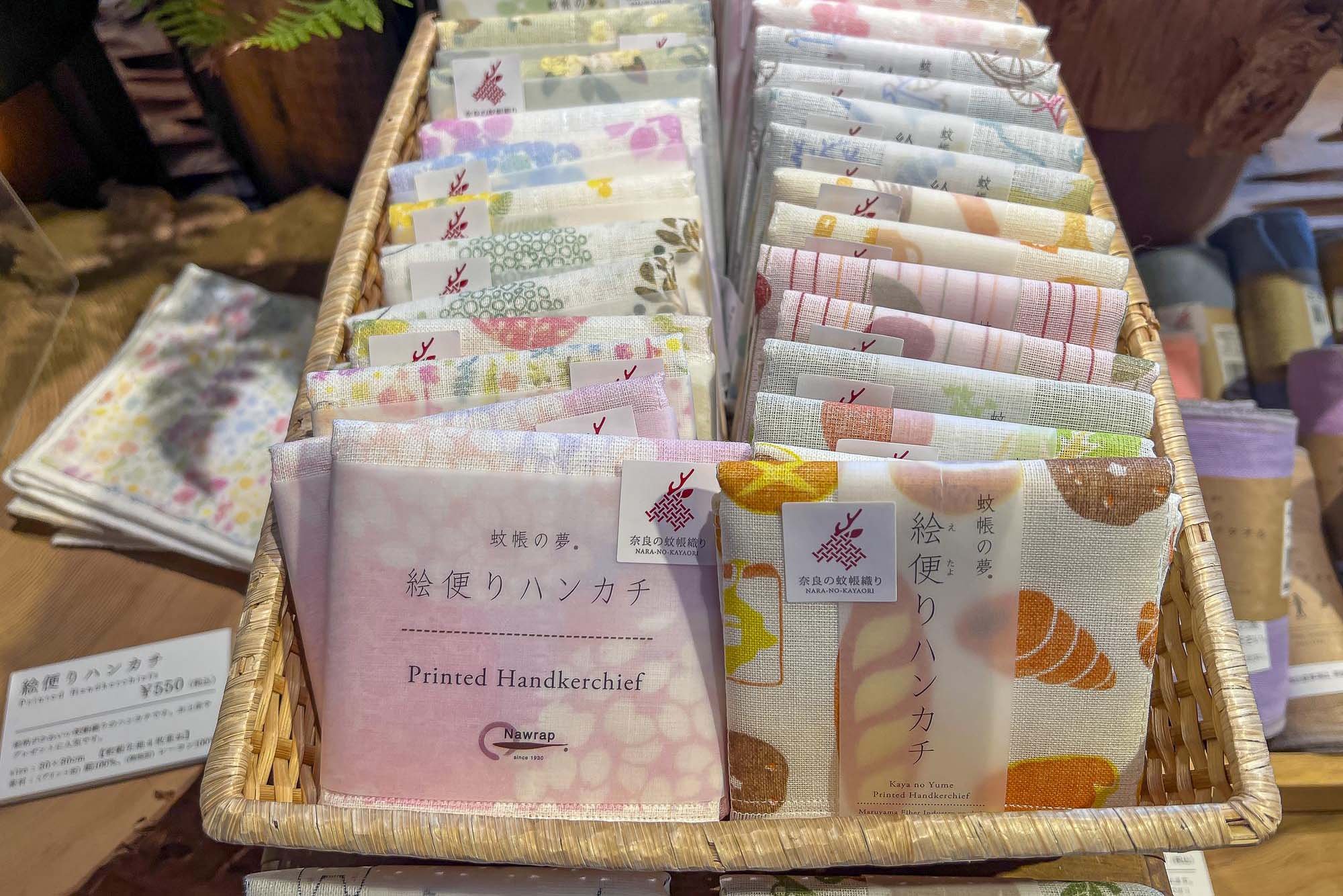 A woven basket containing folded printed handkerchiefs with floral and pattern designs, labeled with tags displaying Japanese characters and the words 'Printed Handkerchief' in English.