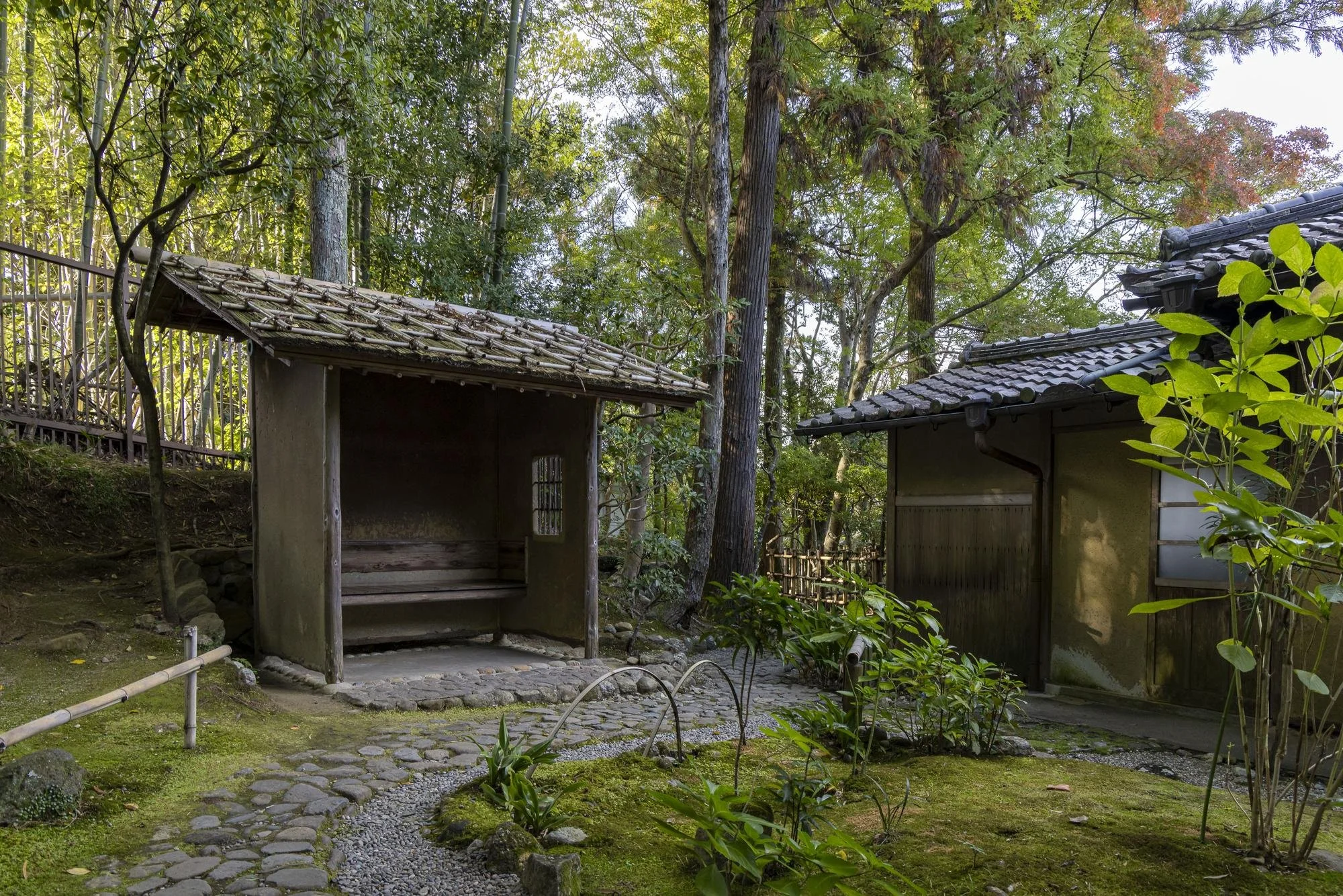 Traditional Japanese garden with two small wooden structures, surrounded by green trees and plants, a cobblestone pathway, and mossy ground.