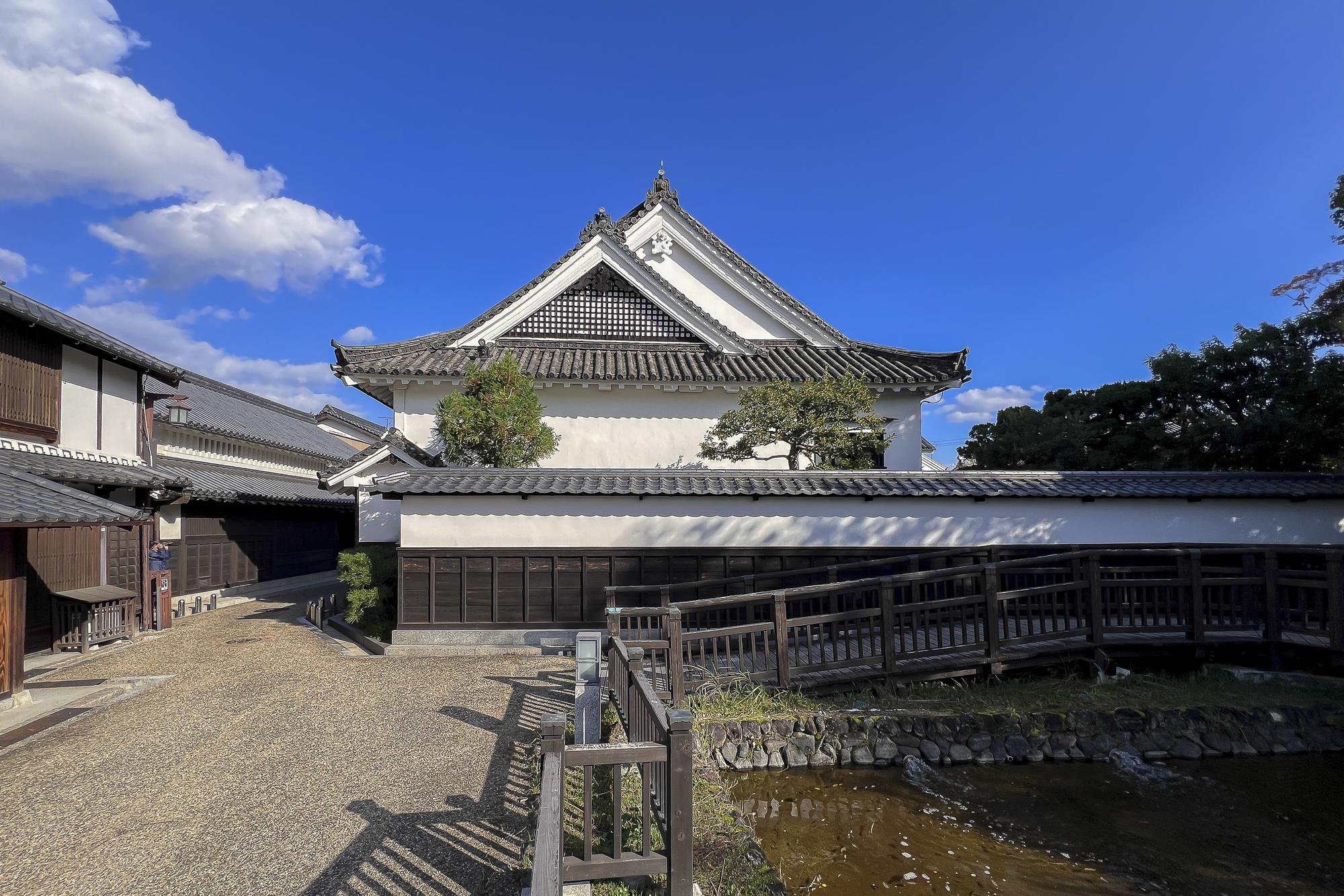 Traditional Japanese building with white walls and gray tiled roof under a blue sky with clouds, outdoor pathway, small water feature in foreground, and trees around the building.