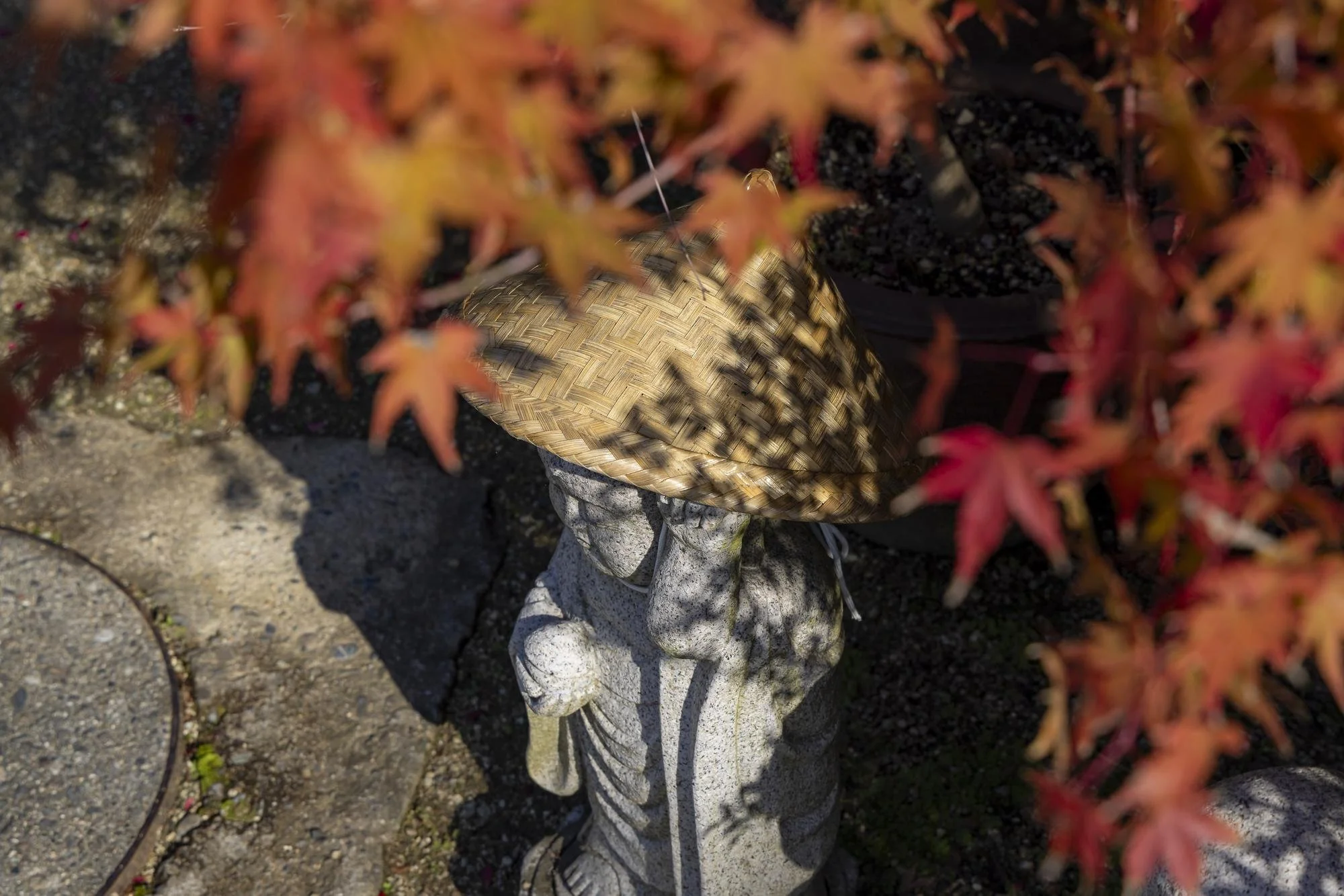 A stone carving of a person holding a large straw hat, partially obscured by red and orange leaves from a plant