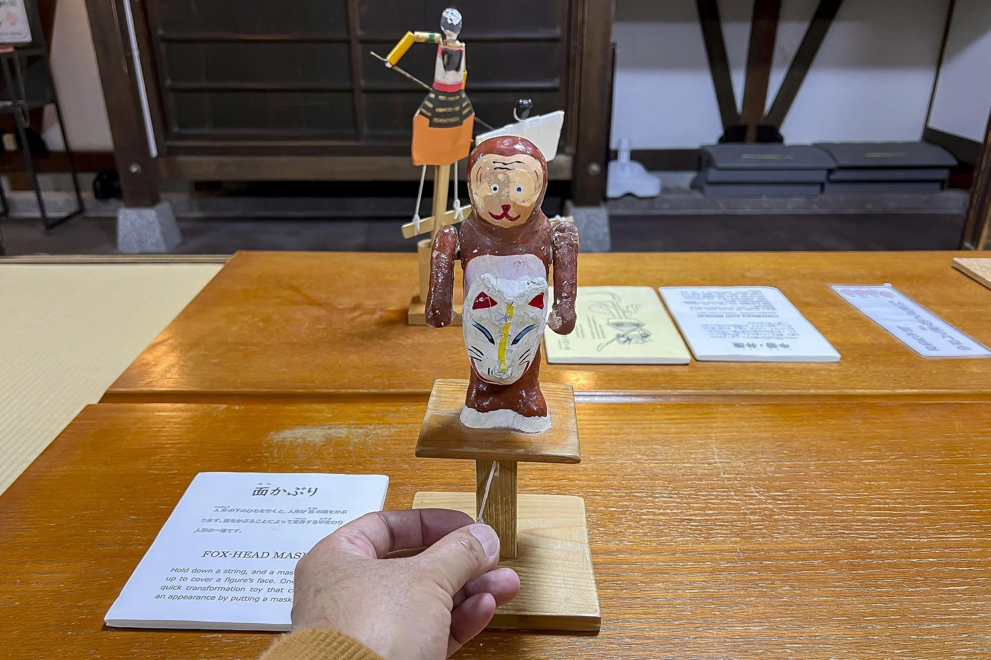 A small, handcrafted figure resembling a human holding a mask in front of face with intricate designs, mounted on a wooden stand, displayed on a wooden table.