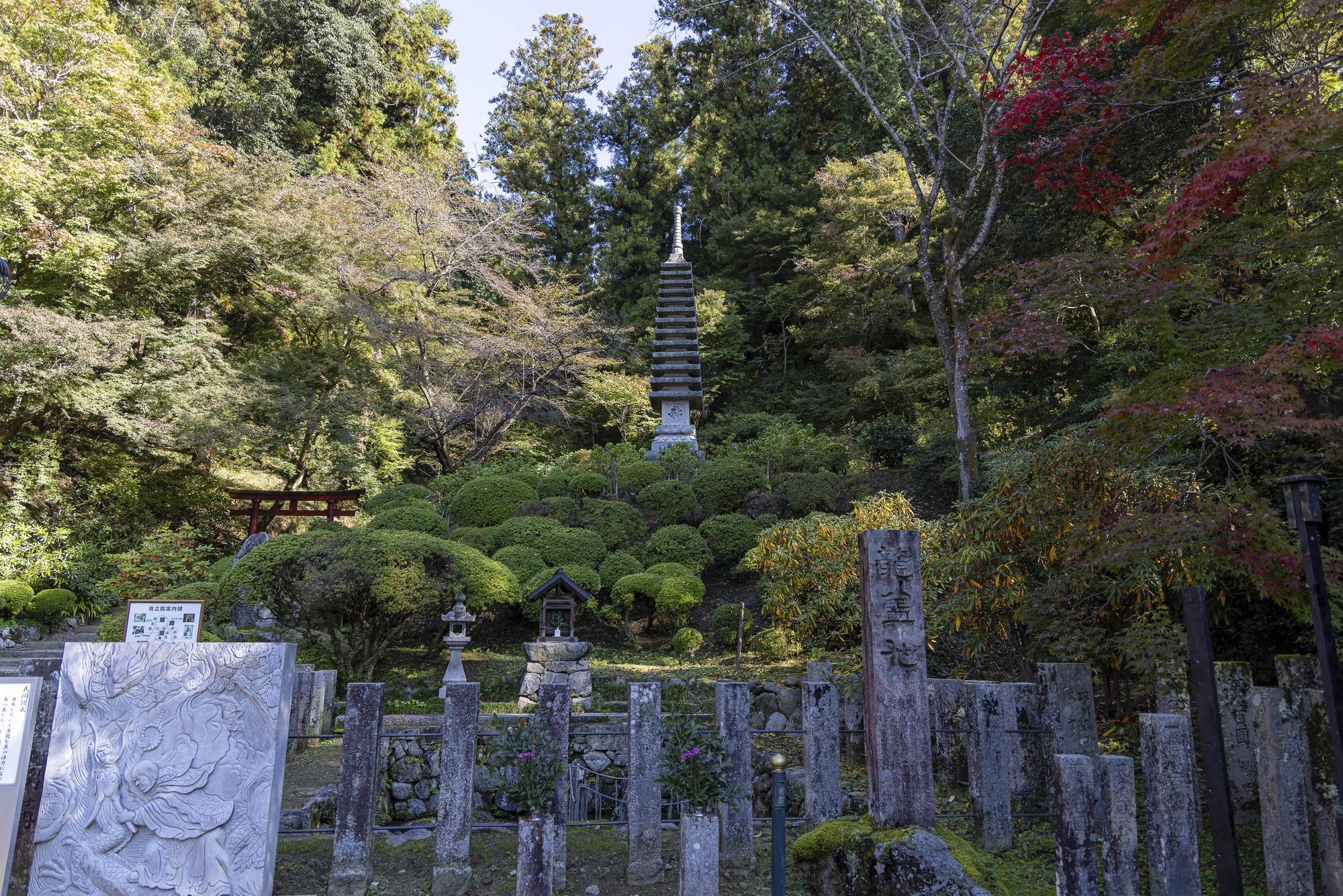 View of a temple site with stone markers, lanterns, and a small shrine, surrounded by lush green trees and bushes, with a tall stone pagoda in the background, in a forested area.