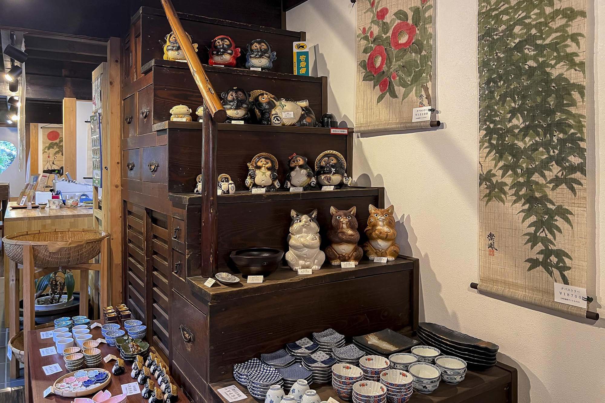 A display of various Japanese-themed ceramic figurines and dishware in a store. The ceramic figurines include tanuki and cat figures, while the dishware includes small bowls and plates with traditional patterns. There are hanging scrolls with Japanes