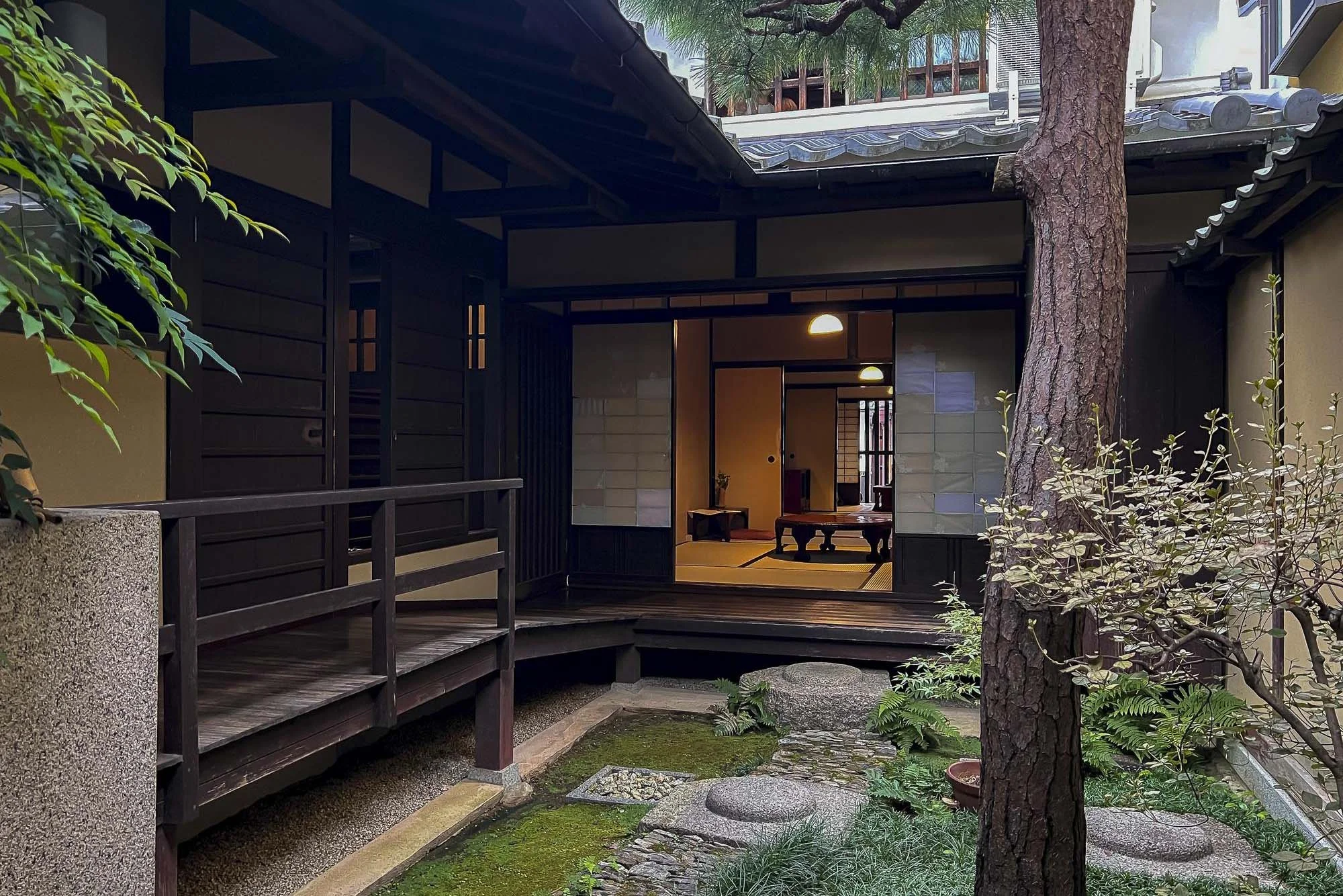 Traditional Japanese-style house with an open sliding door leading into a tatami room, surrounded by a small garden with trees and rocks.