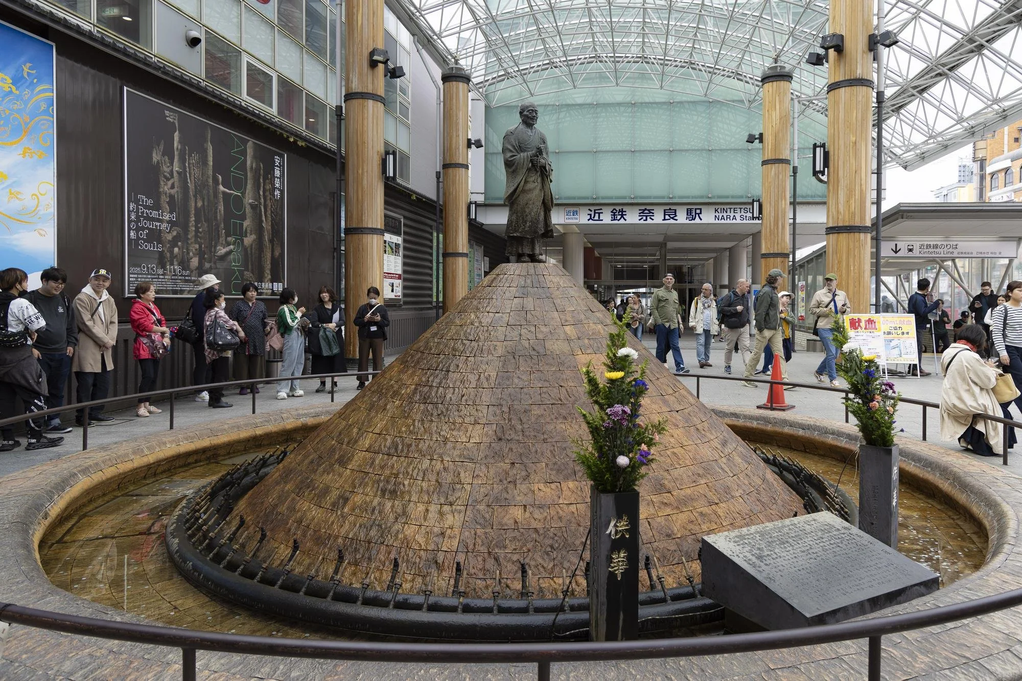 A fountain with a conical shape and a statue of a man at the top, located outside a train station with a crowd of people waiting in line and walking nearby.