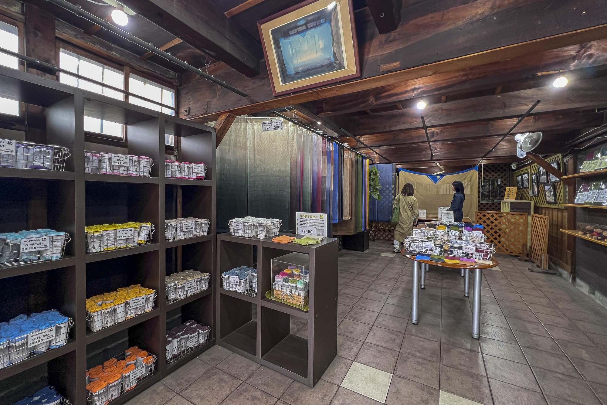 Interior of a store displaying colorful fabrics hanging vertically and jars of yarn or thread arranged on shelves and a table.
