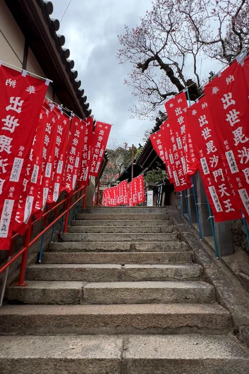 Stone steps leading up to a Japanese temple or shrine, flanked by red banners with white Japanese characters, and a leafless tree in the background under an overcast sky.