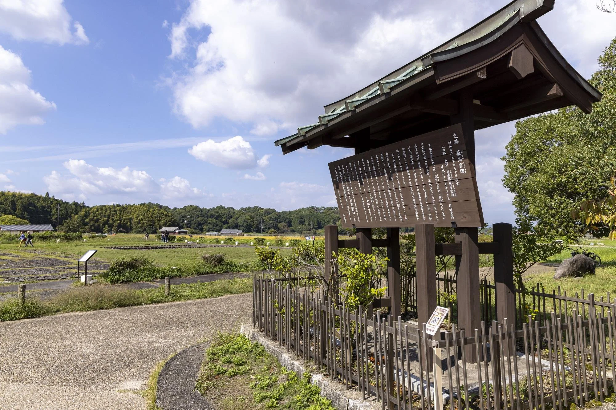Asuka Palace Site: A traditional Japanese wooden sign with Japanese text, surrounded by a small fence, on a tile and concrete walkway with greenery. In the background, there are fields, a few small buildings, trees, and a group of people walking on t