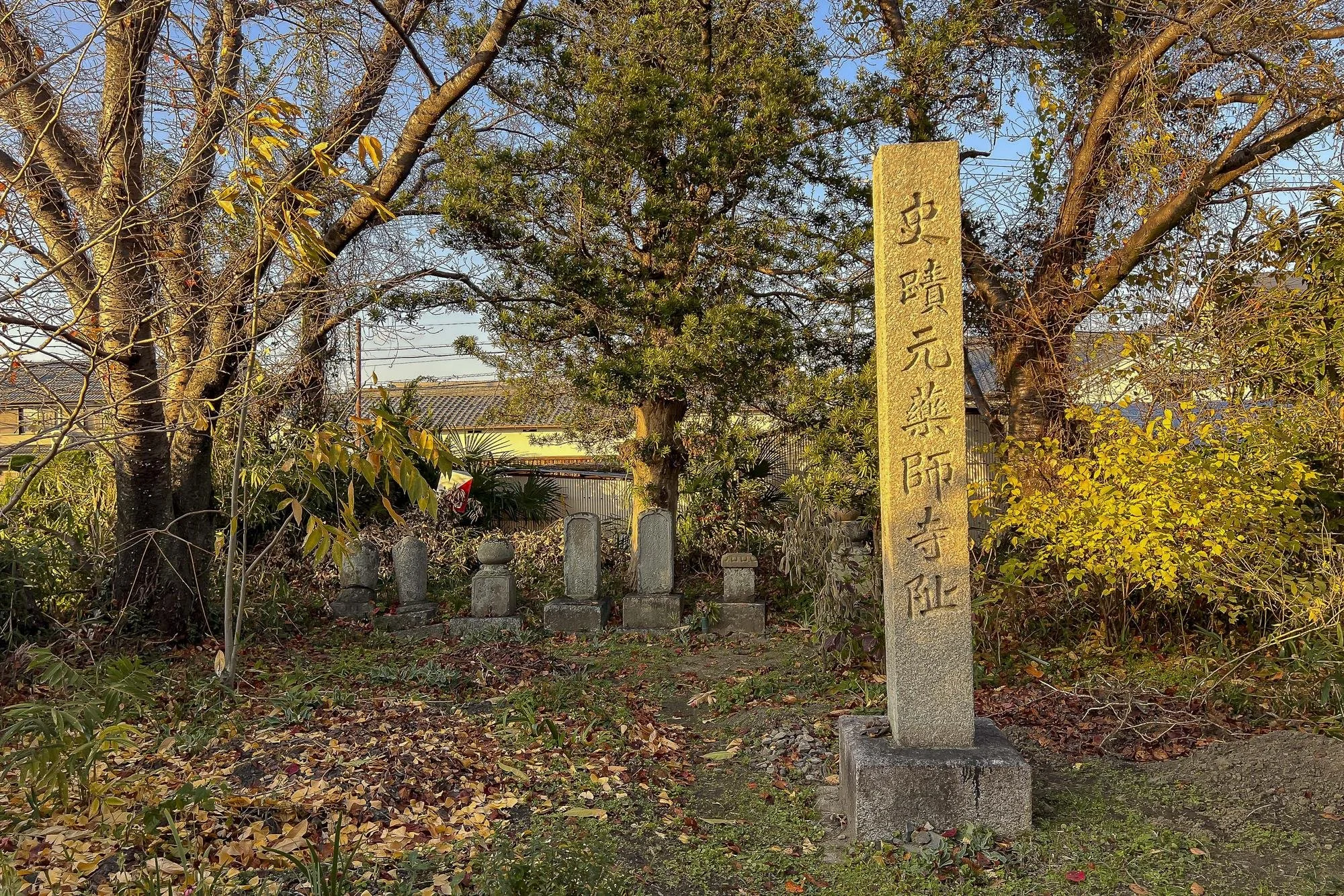 A small traditional Japanese sacred site with stone markers and inscriptions, surrounded by trees and fallen autumn leaves.