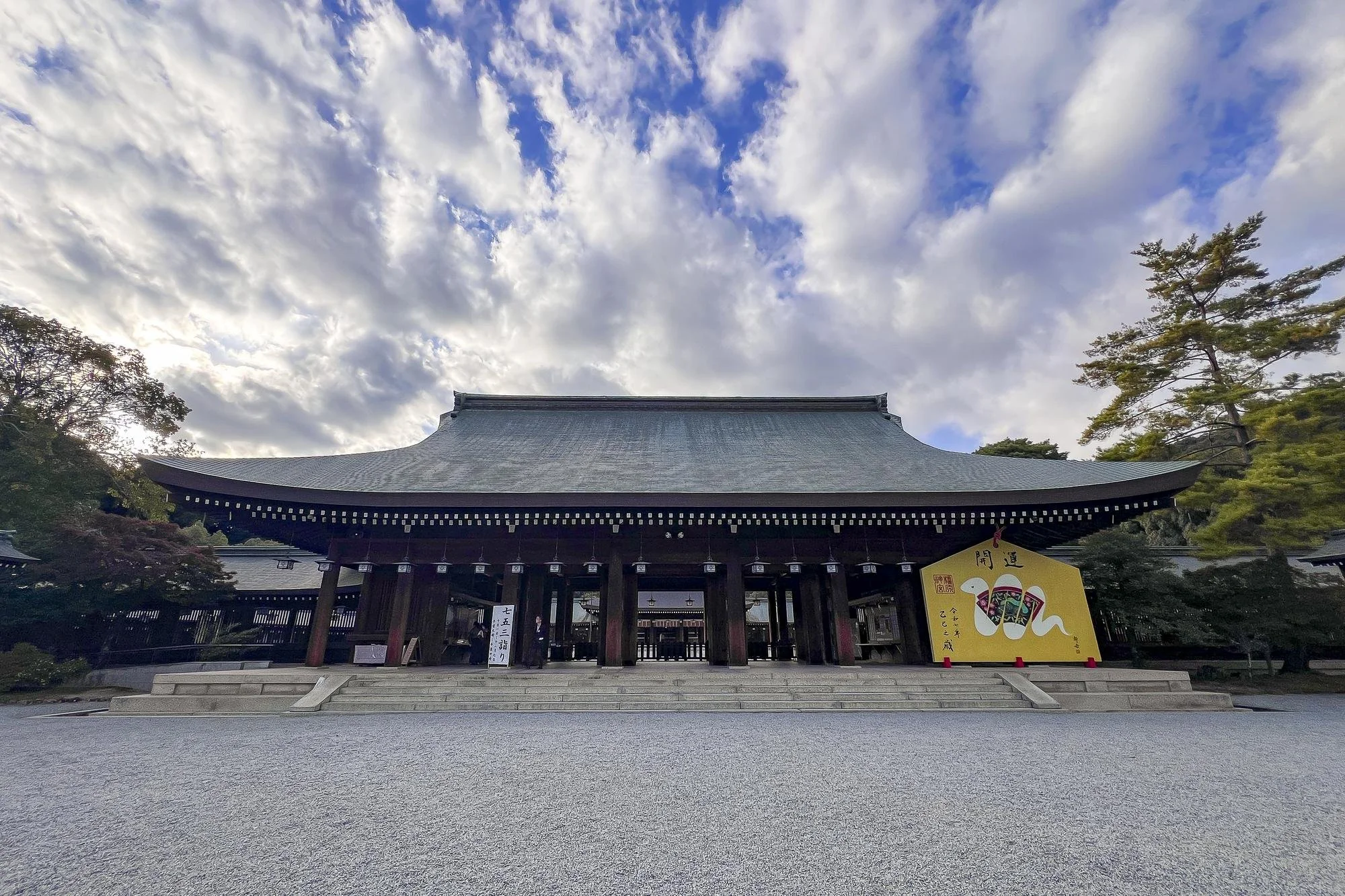 Traditional Japanese shrine with wooden gates and a tiled roof, surrounded by trees and a gravel courtyard, under a partly cloudy sky.