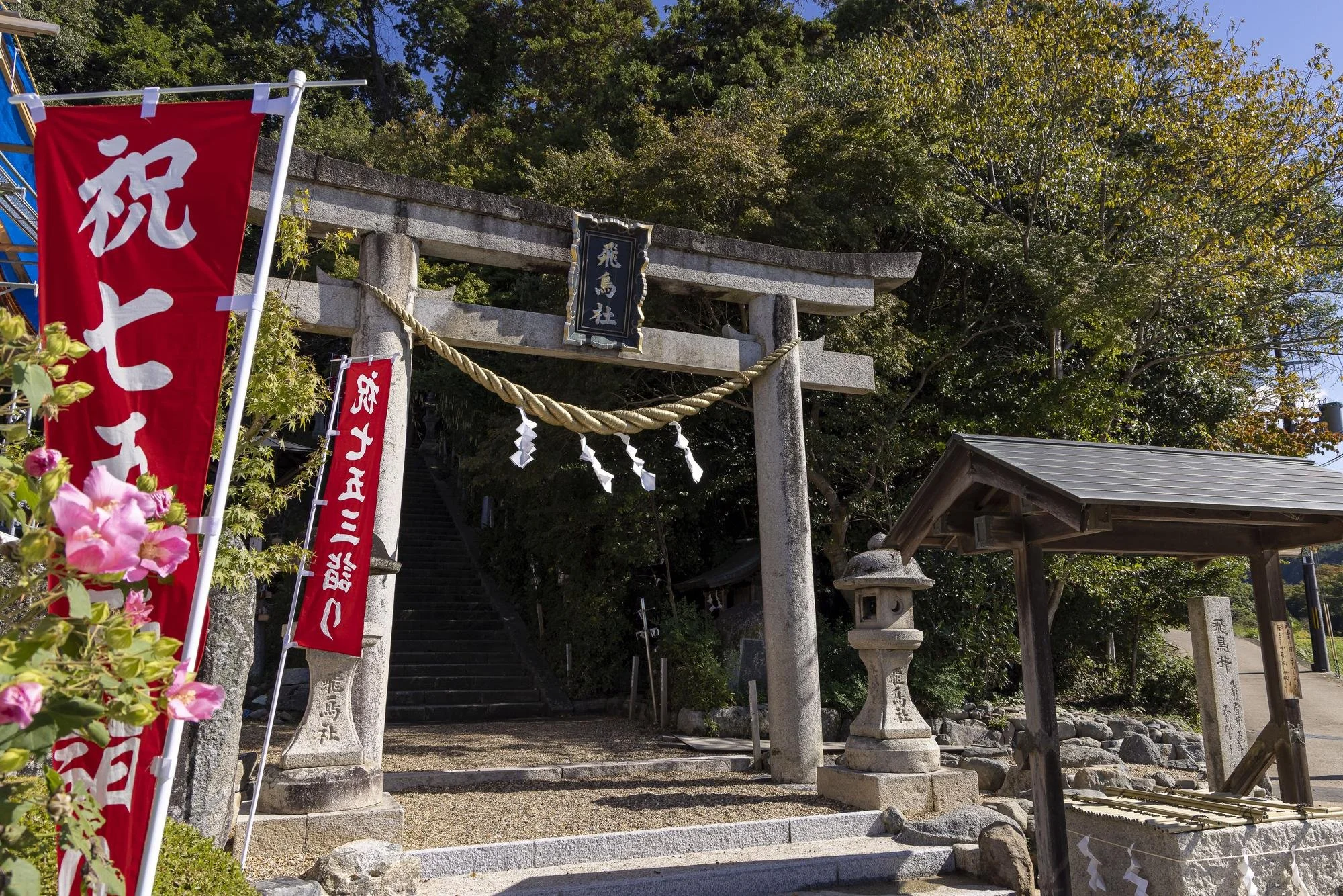 Stone torii gate at the entrance of a Japanese shrine with banners, lantern, and stairs leading up into a wooded area.