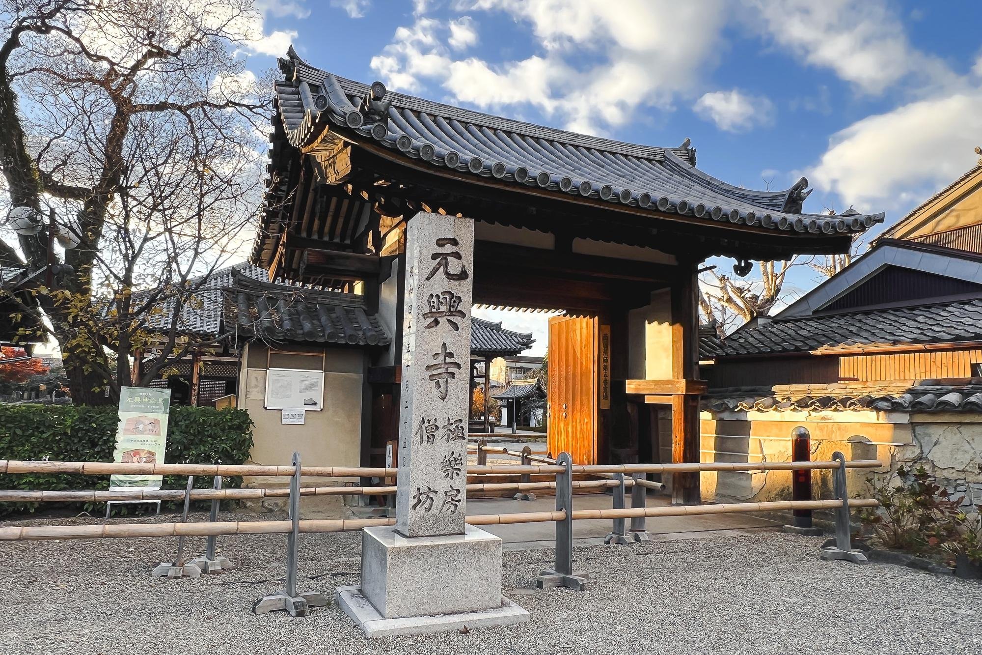 Traditional Japanese temple gate with wooden architecture and stone pillar, surrounded by trees and a gravel path, under a partly cloudy sky.