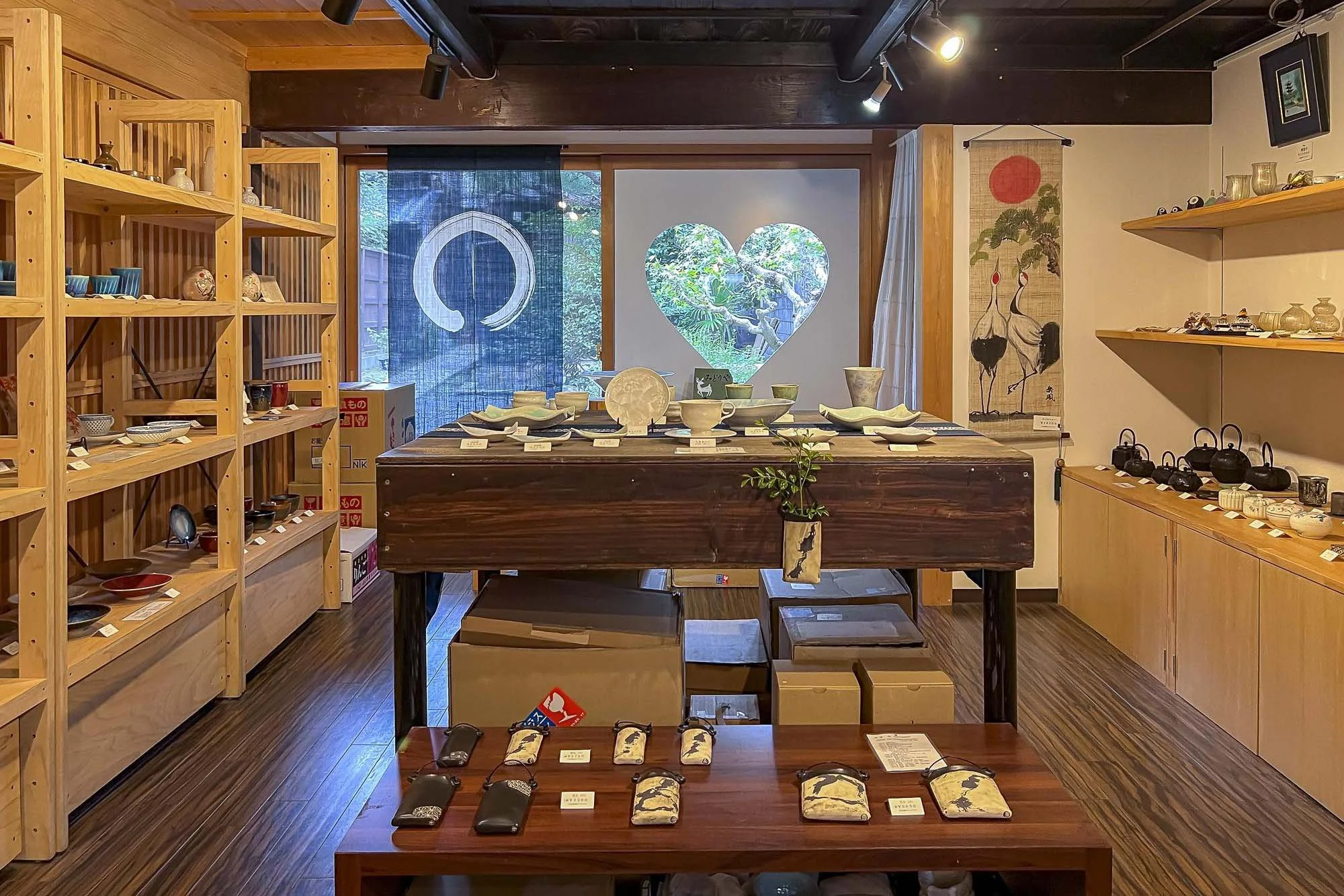 Interior of a store displaying various ceramic and pottery items, with shelves on the left and right, and a wooden table in the center showcasing bowls, pots, and accessories. A heart-shaped window and traditional Japanese hanging scroll are visible in the background.