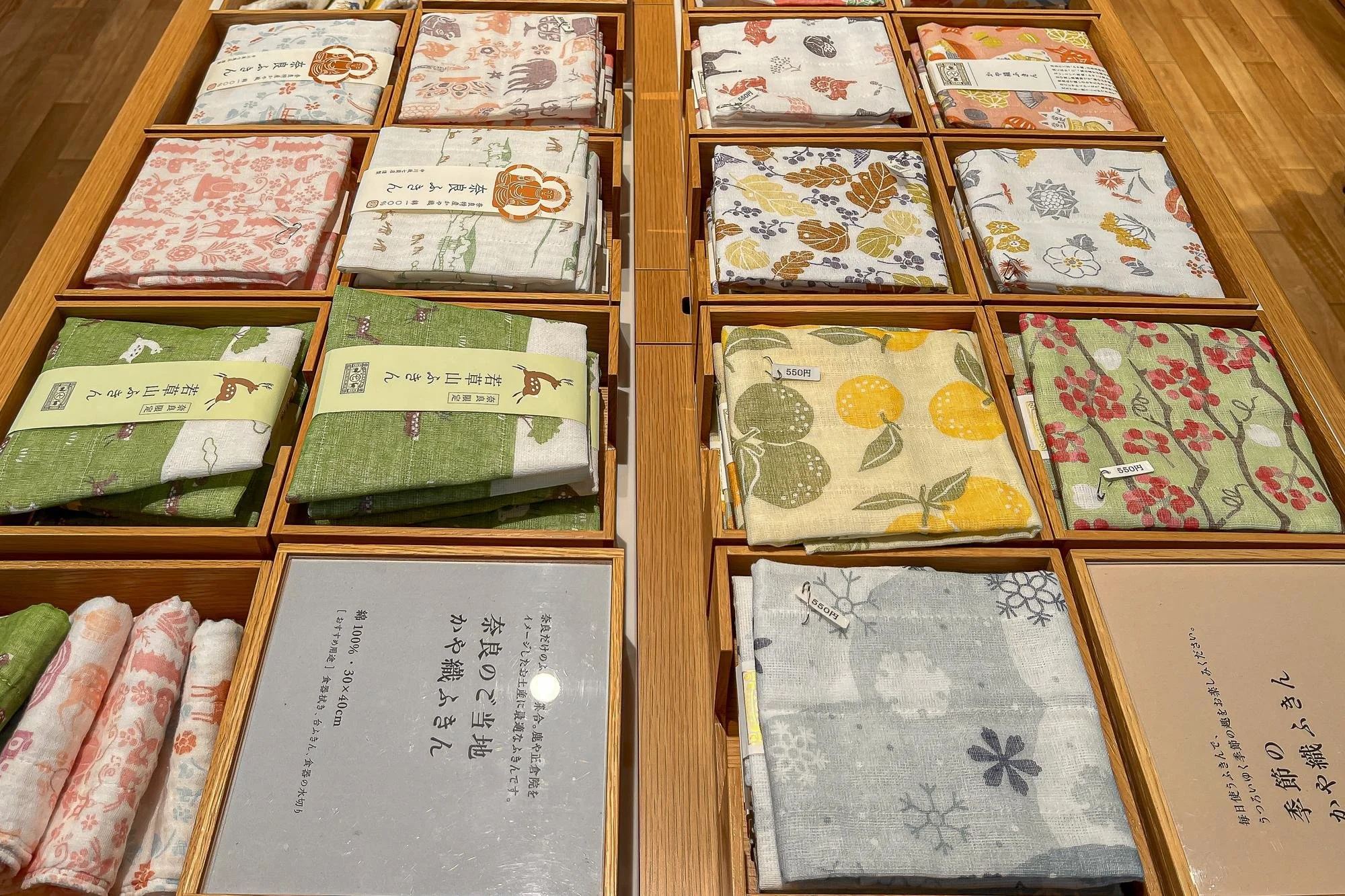 Display of folded patterned cloth napkins in wooden trays on a wooden surface at a store, with some labeled with price tags.