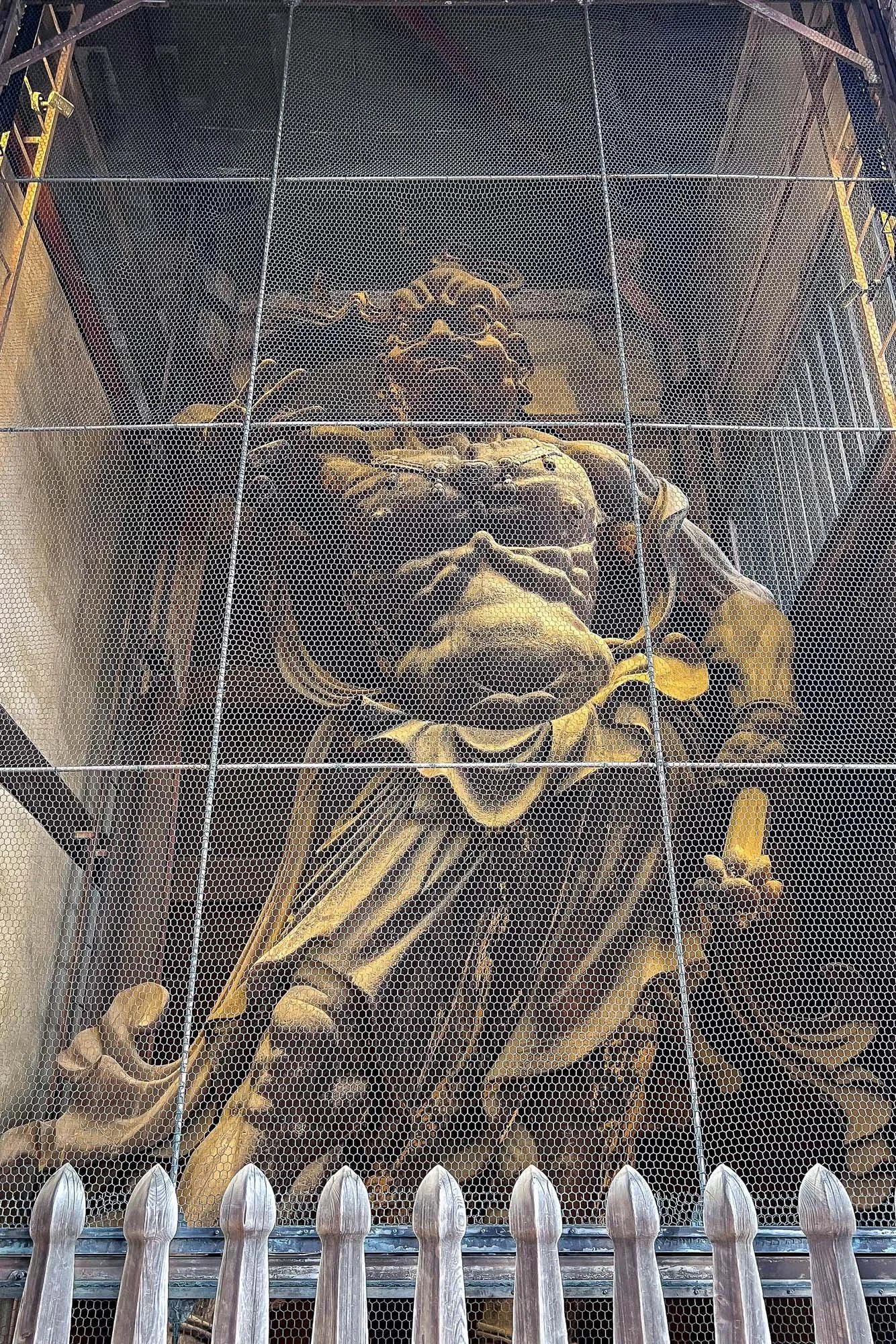 A large stone sculpture of a religious figure, possibly a saint or angel, behind a protective wire mesh at a historical or religious site.