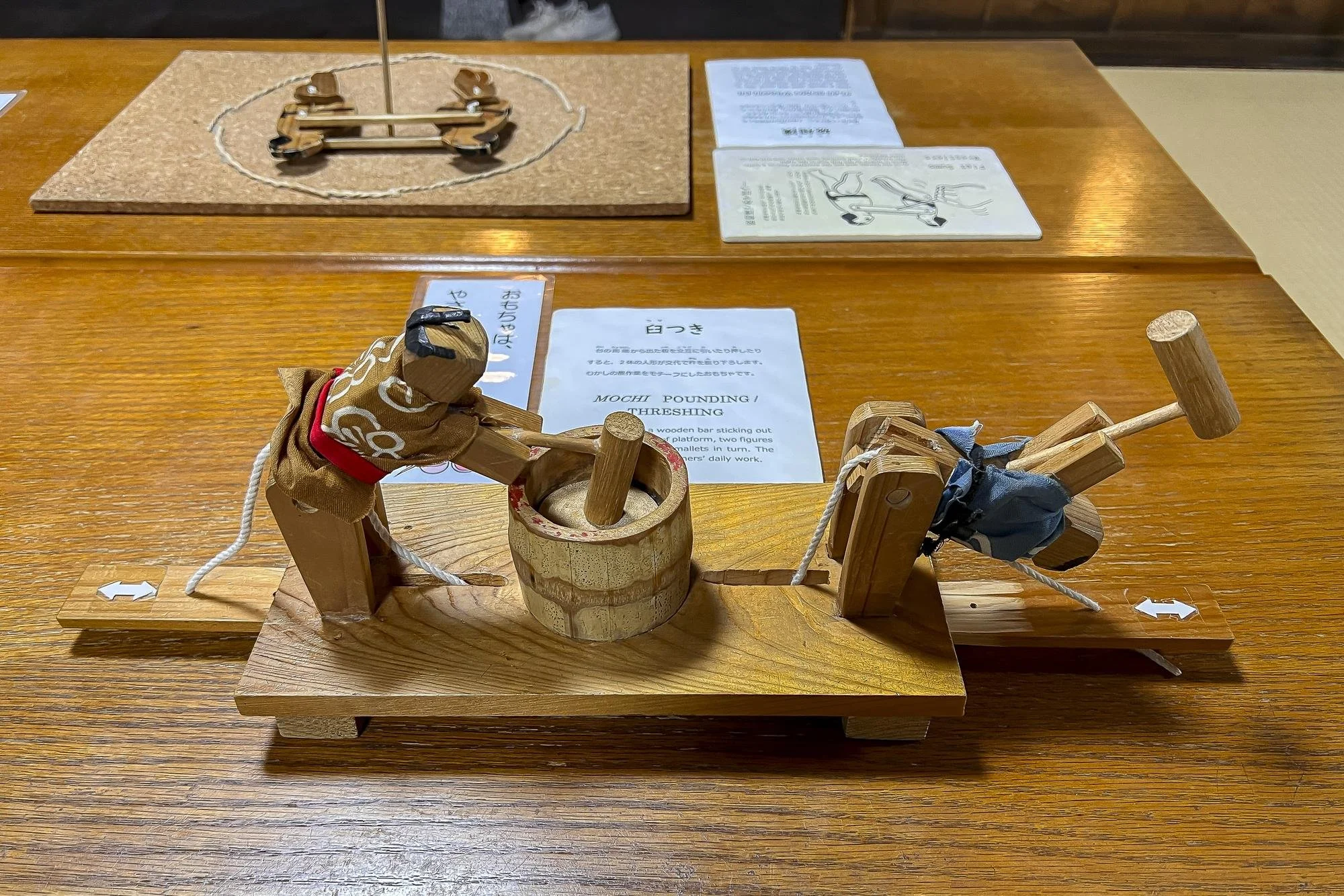 Wooden toy noisemaker featuring two figures on a platform, pulling ropes connected to a drum in the middle, on a wooden table with printed instructions in the background.