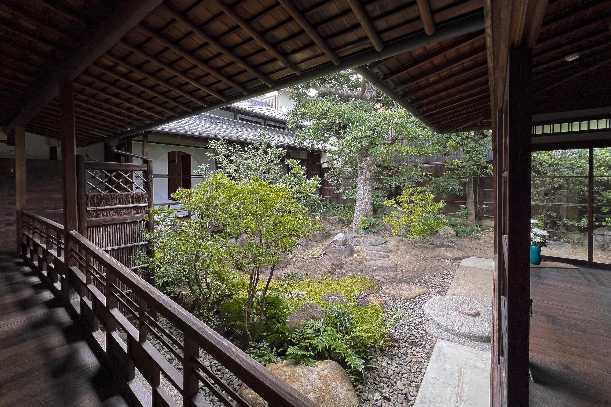 Traditional Japanese-style courtyard garden viewed from a covered wooden porch with plants, rocks, and a tree