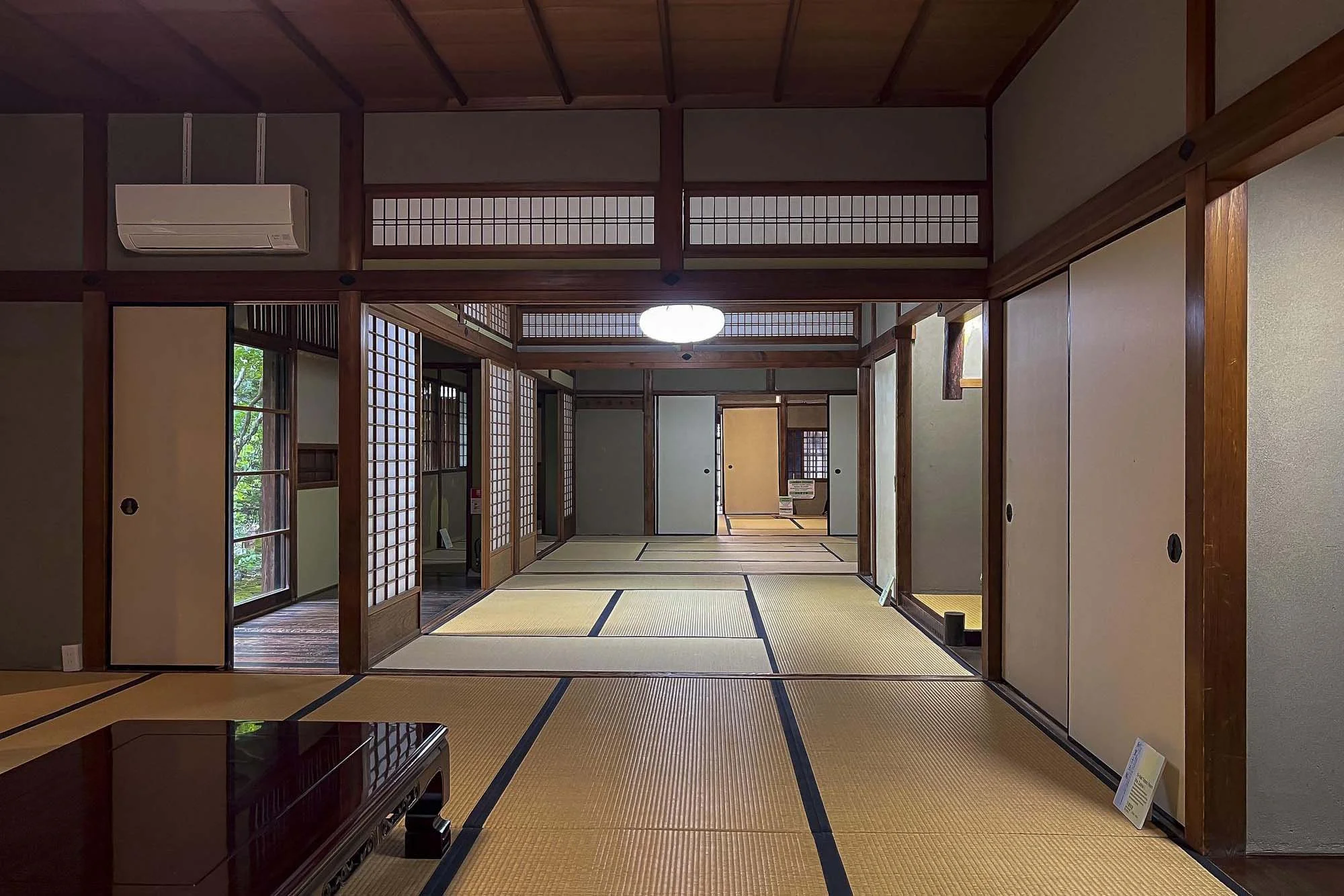 Interior of a traditional Japanese room with tatami mats, sliding doors, and wooden accents, overlooking a garden with trees.