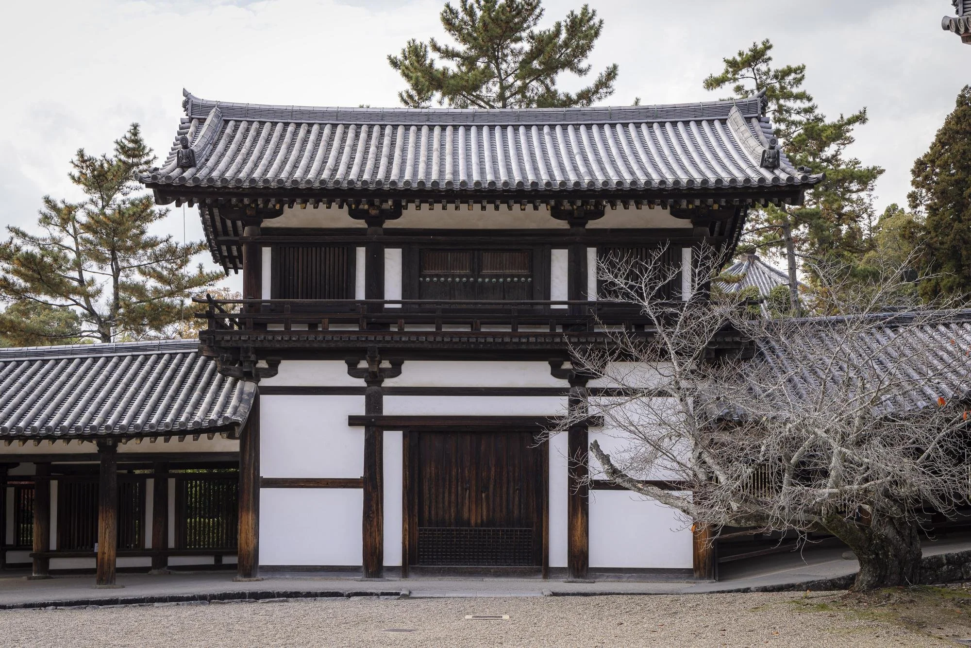 Traditional Japanese building with wooden and white walls, a tiled roof, trees, and a gravel path in front.