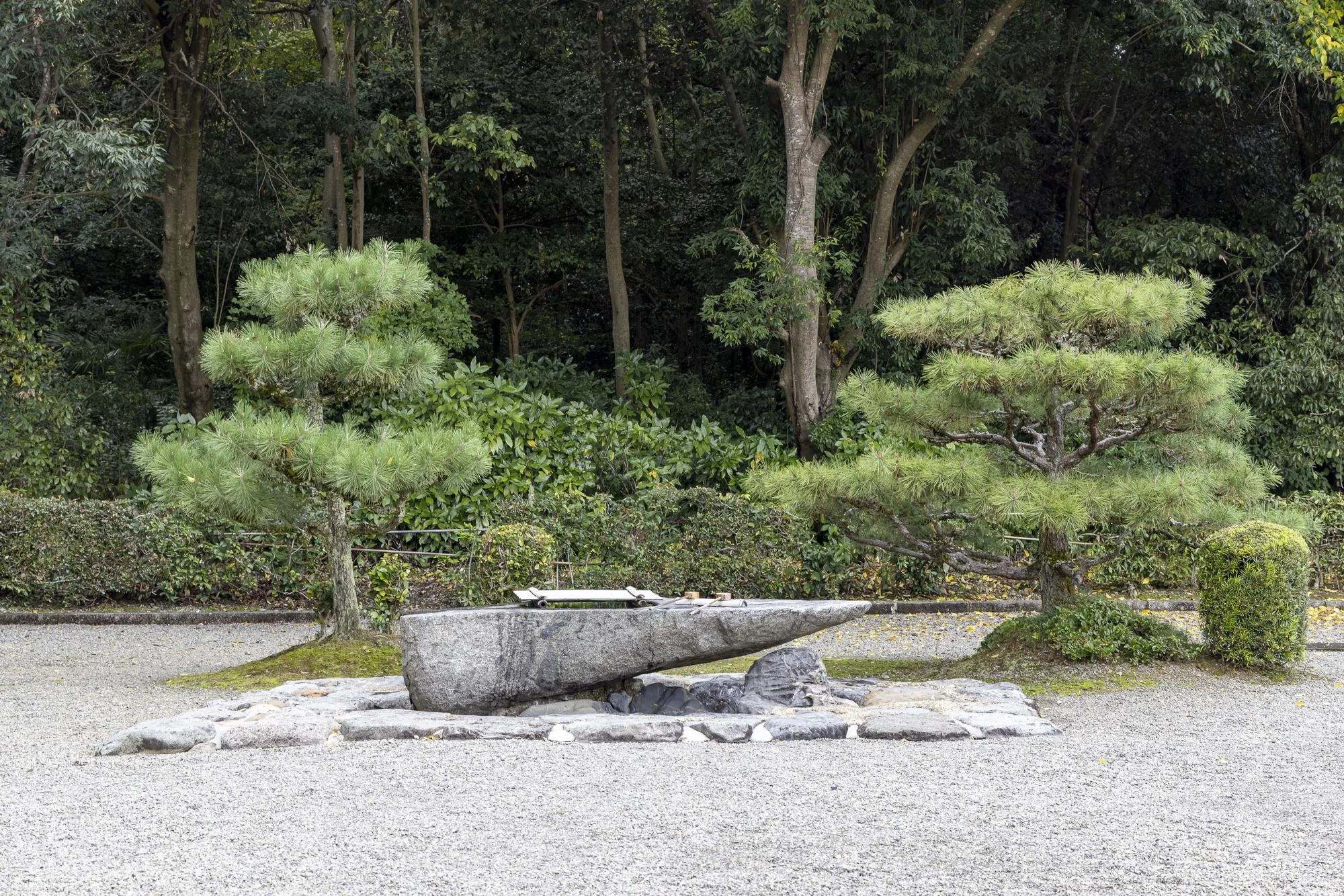 A traditional Japanese rock garden featuring a large, horizontally placed gray stone with a smaller stone beneath it, surrounded by neatly raked gravel, two pruned pine trees, and lush green bushes set against a backdrop of tall trees.