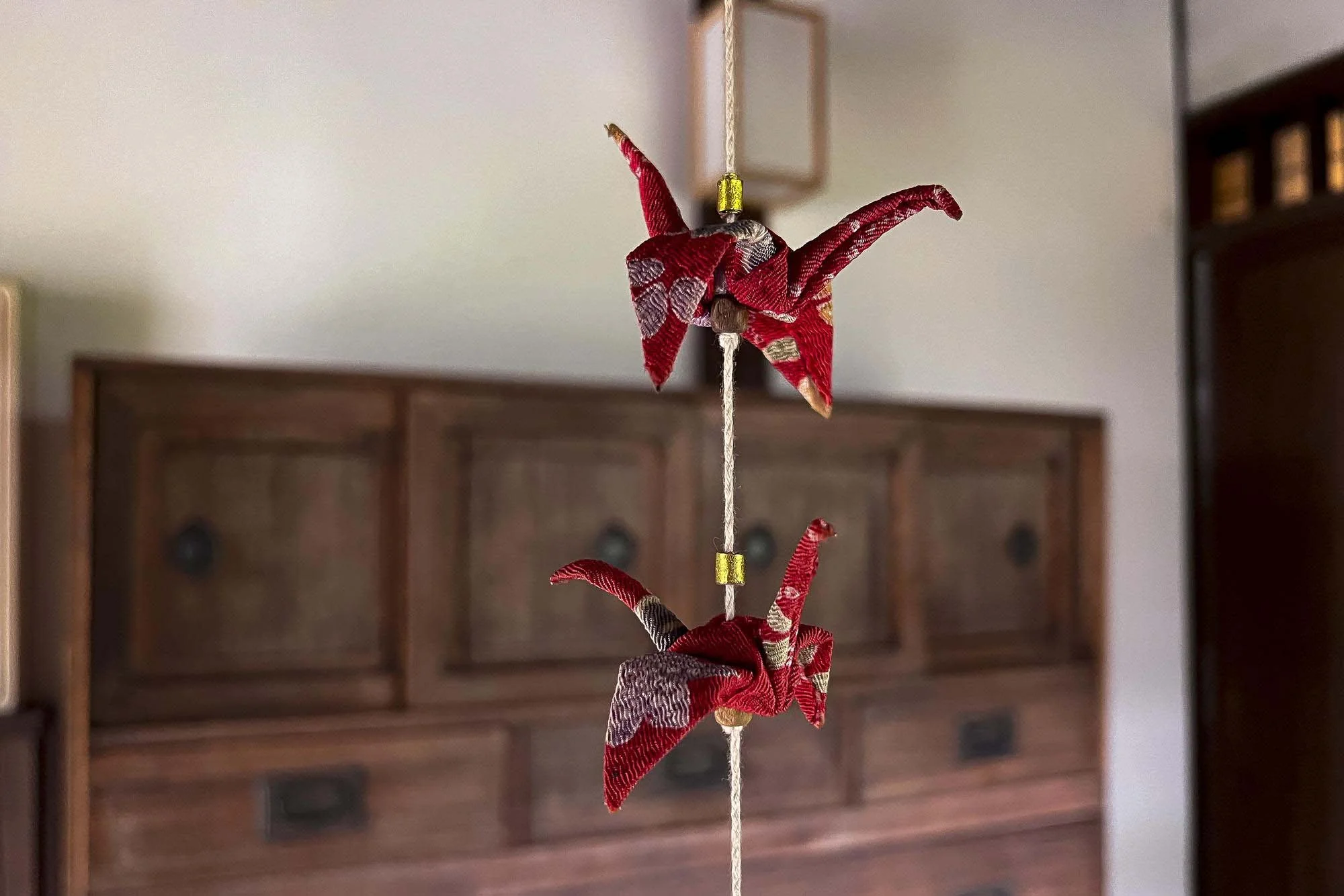 Red fabric origami cranes hanging on string indoors with a wooden cabinet in the background.