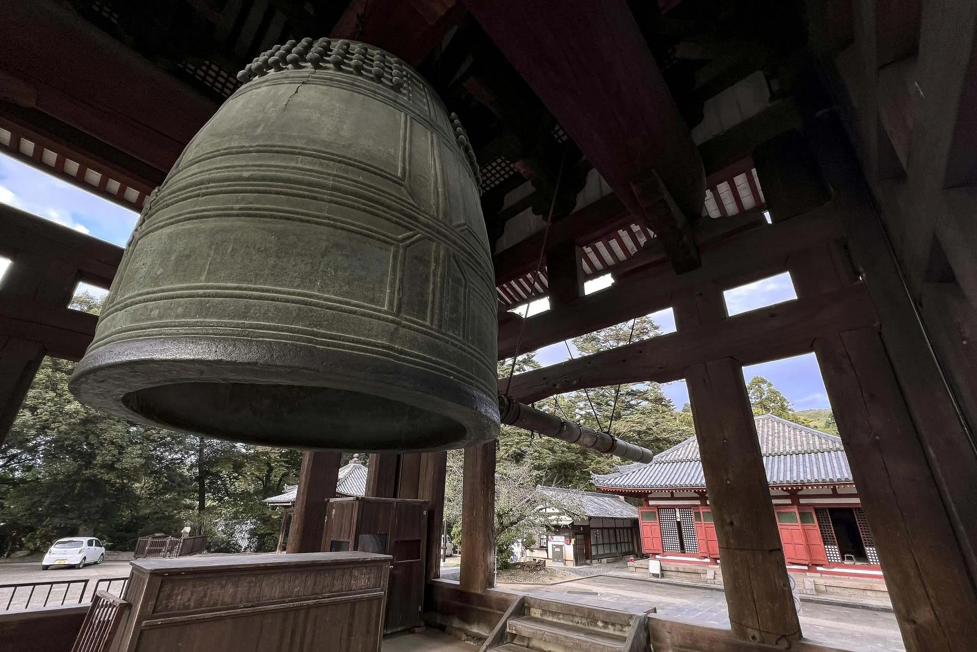 Large bronze bell hanging inside a traditional Japanese temple structure with wooden beams and open sides, showing temple buildings and trees outside.