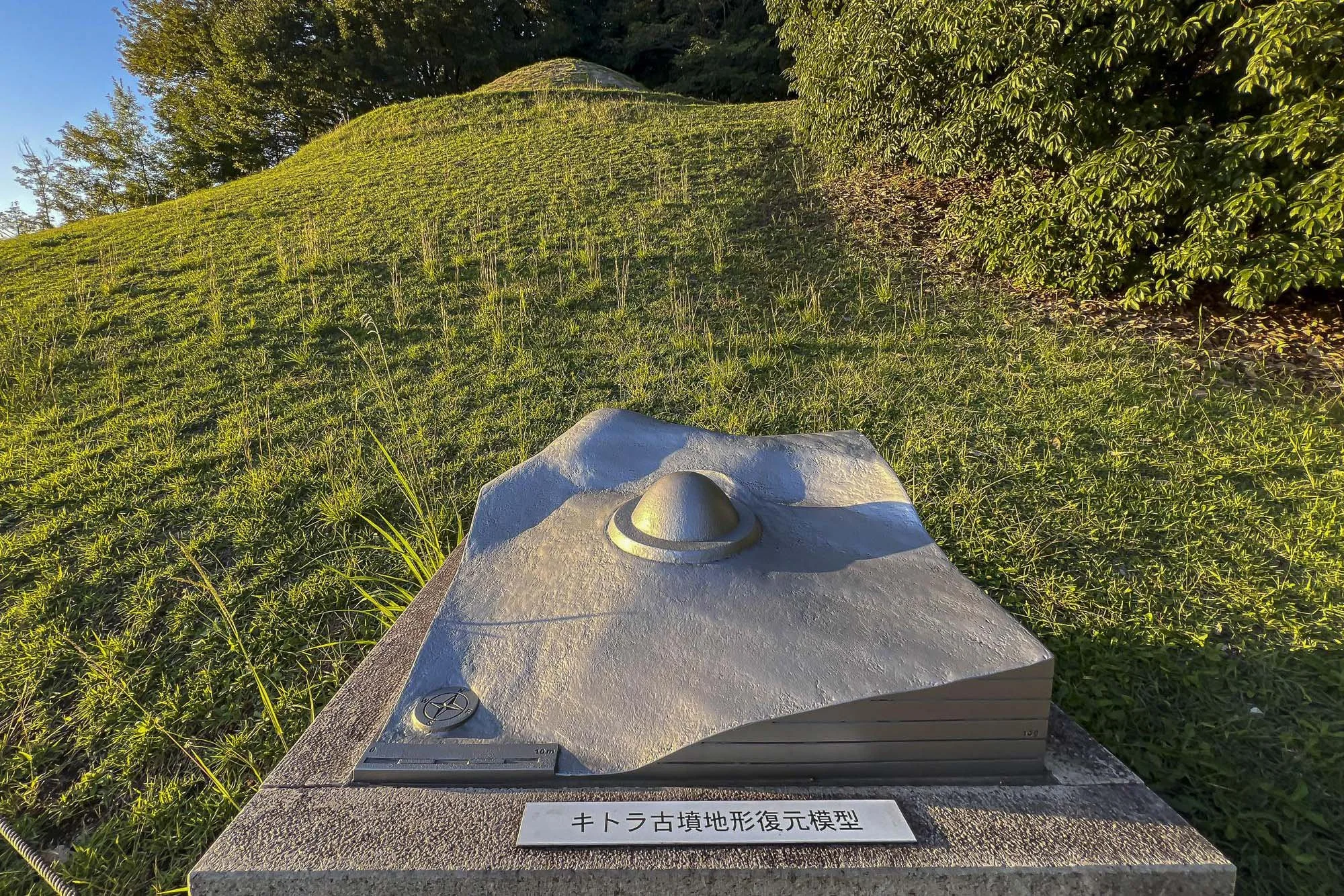 A topographic model of an archaeological site with a grassy hill, trees in the background, and a metallic model of the landscape placed on a stone pedestal. The pedestal has a label in Japanese characters.