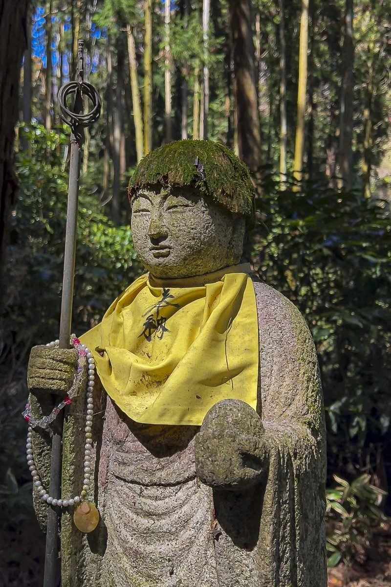 Stone statue of a Buddhist monk with mossy hair, wearing a yellow cloth and holding a staff with prayer beads, in a forest setting.