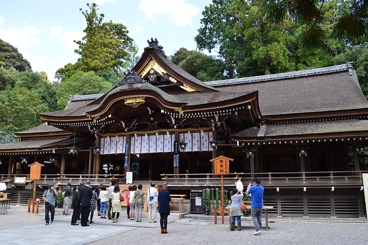 A traditional Japanese shrine with a richly decorated wooden roof, surrounded by lush green trees. People are gathered in front of the shrine, some taking photos and others praying or observing.