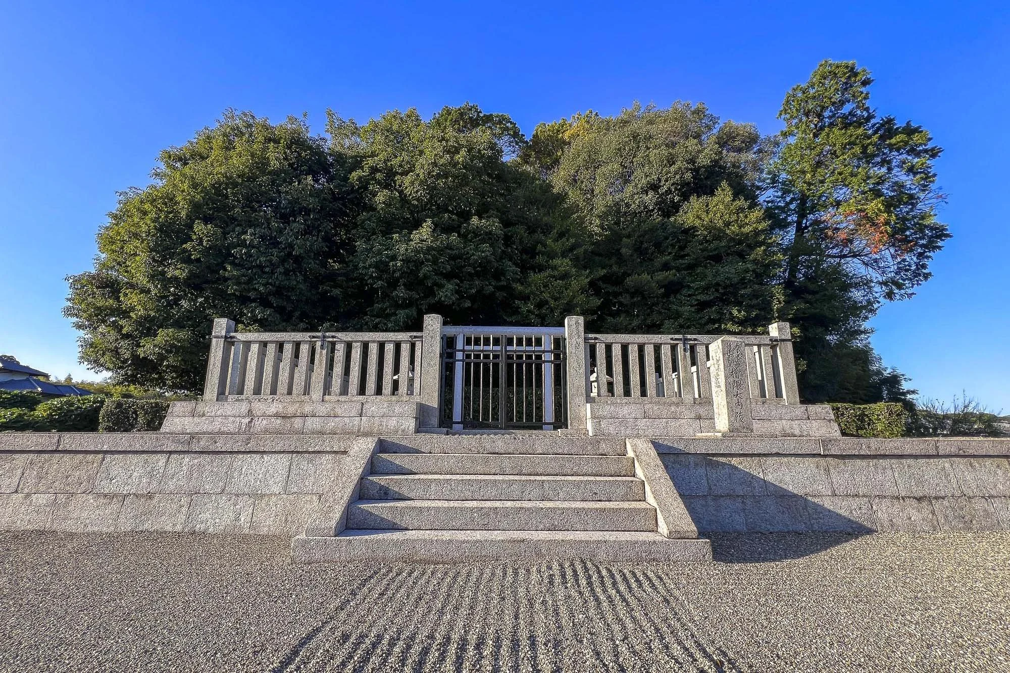 Mounded Tomb of Emperor Temmu and Empress Jito: Stone steps leading up to a small gate with a wooden fence, with large green trees in the background and a clear blue sky.
