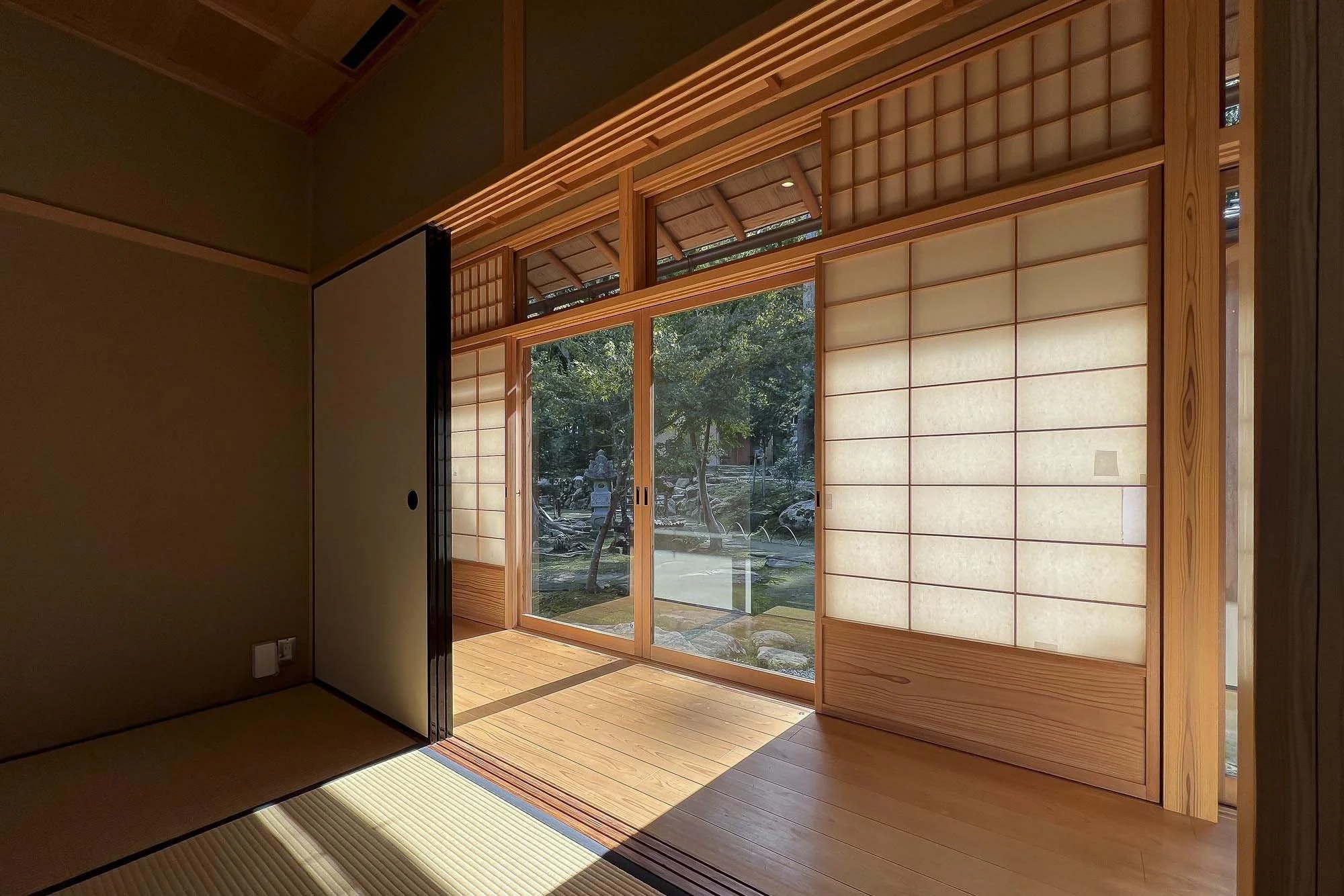 A traditional Japanese room with shoji sliding doors opening to a garden with trees and rocks, sunlight illuminating the wooden floor.