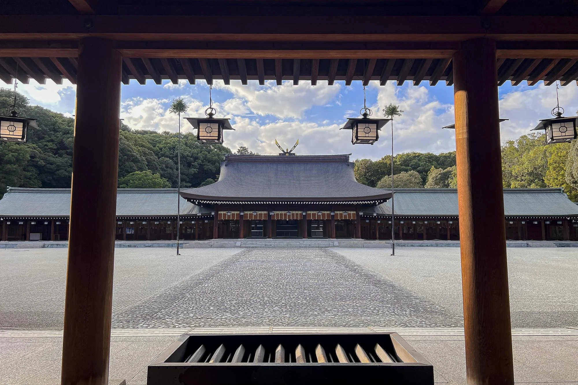 View of a traditional Japanese shrine with a pathway leading to the main hall, framed by wooden pillars and hanging lanterns, surrounded by green trees and a blue sky with clouds.