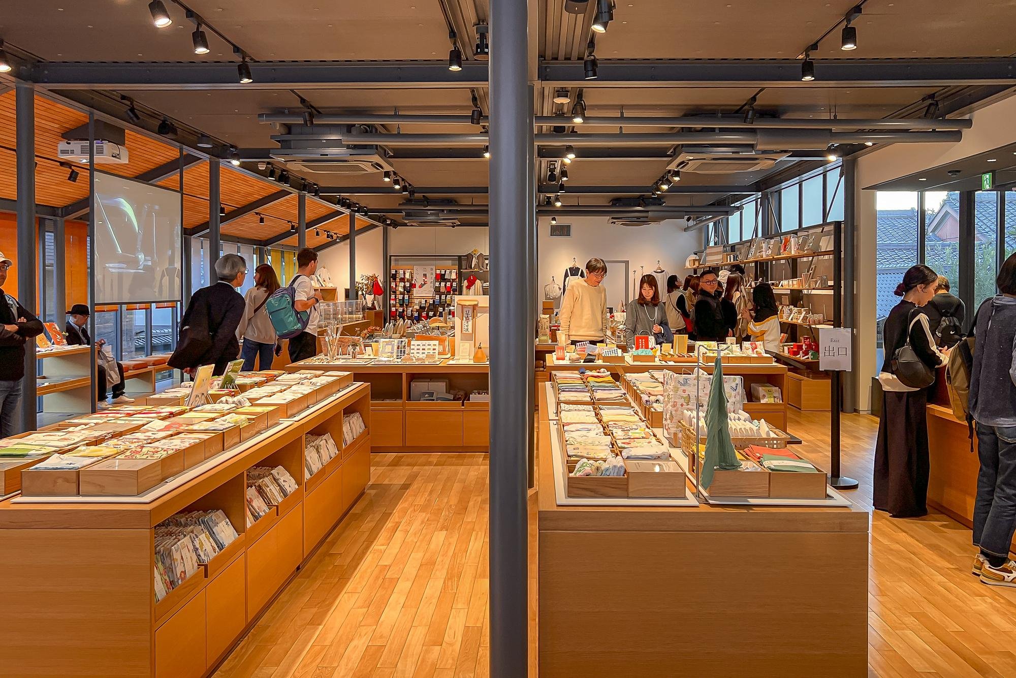 A bookstore with wooden display tables filled with books, magazine, and notebooks. Several customers browse the shelves, and the space is illuminated with black track lighting and large windows allowing natural light.