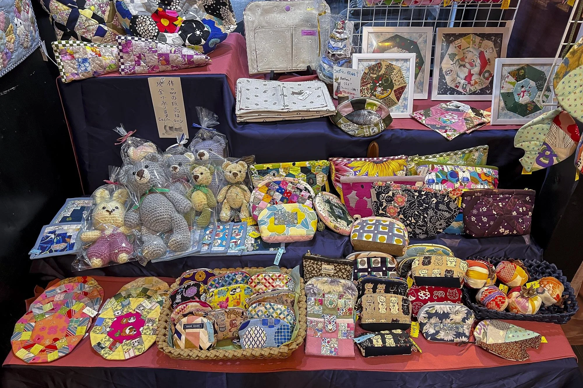 Display of handmade fabric and knit crafts, including bags, pouches, towels, and toys at a craft market stall.