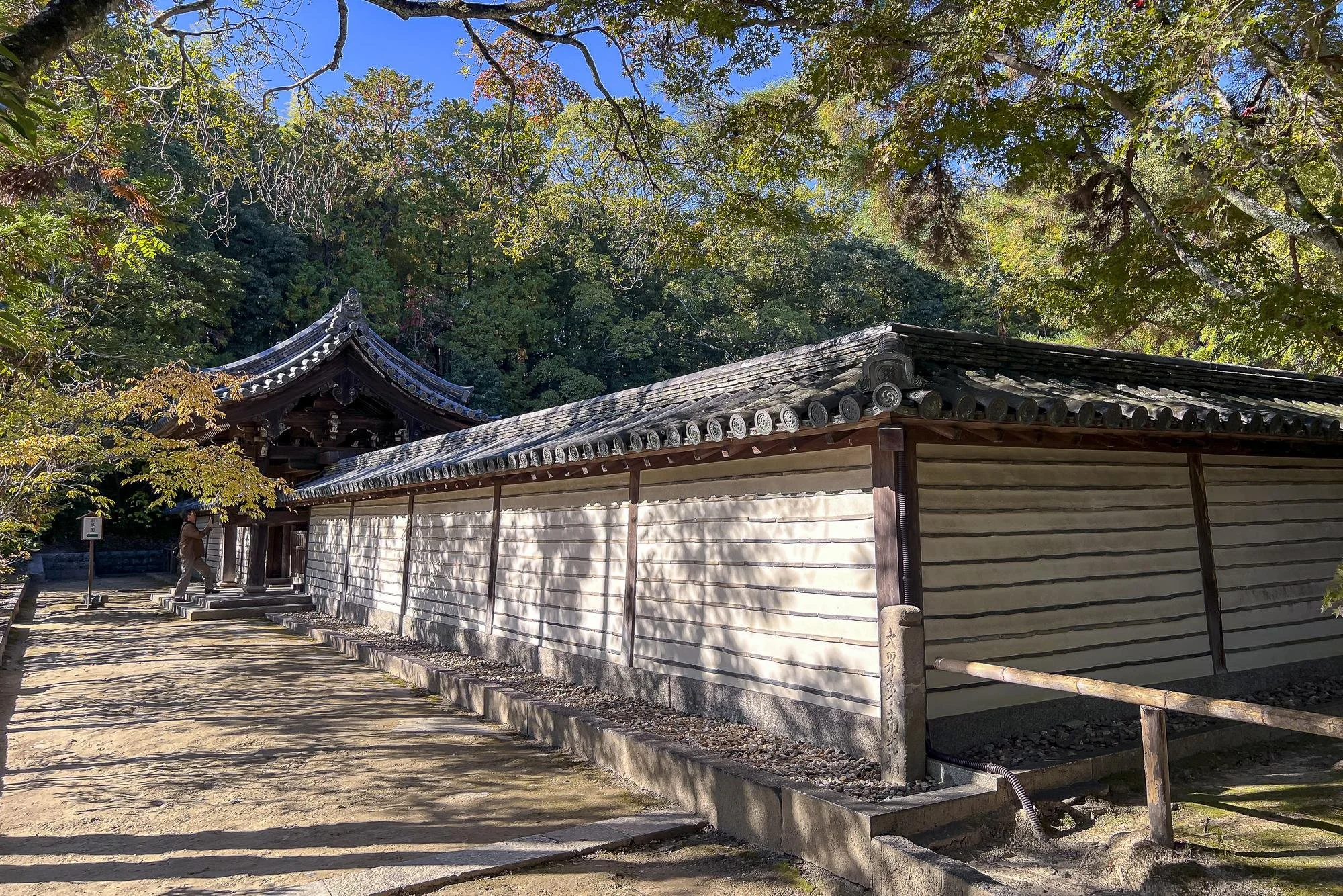 A traditional Japanese building with a tiled roof, wooden walls, and a small entrance. There is a person near the entrance, surrounded by lush green trees under a clear blue sky.
