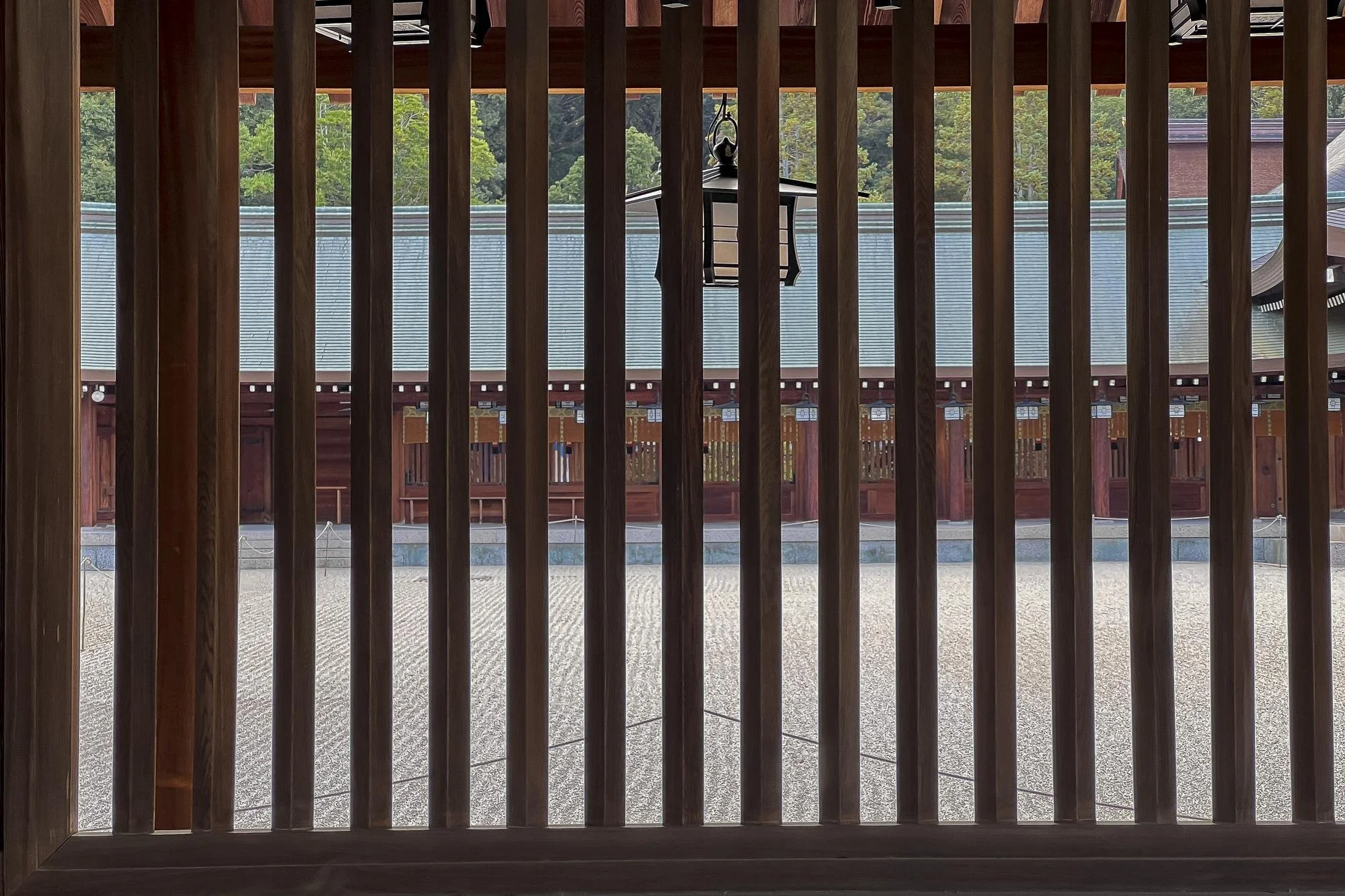 View through wooden vertical slats of a traditional Japanese building with a large lantern hanging in the center, a gravel courtyard, and a row of wooden structures with lanterns in the background.