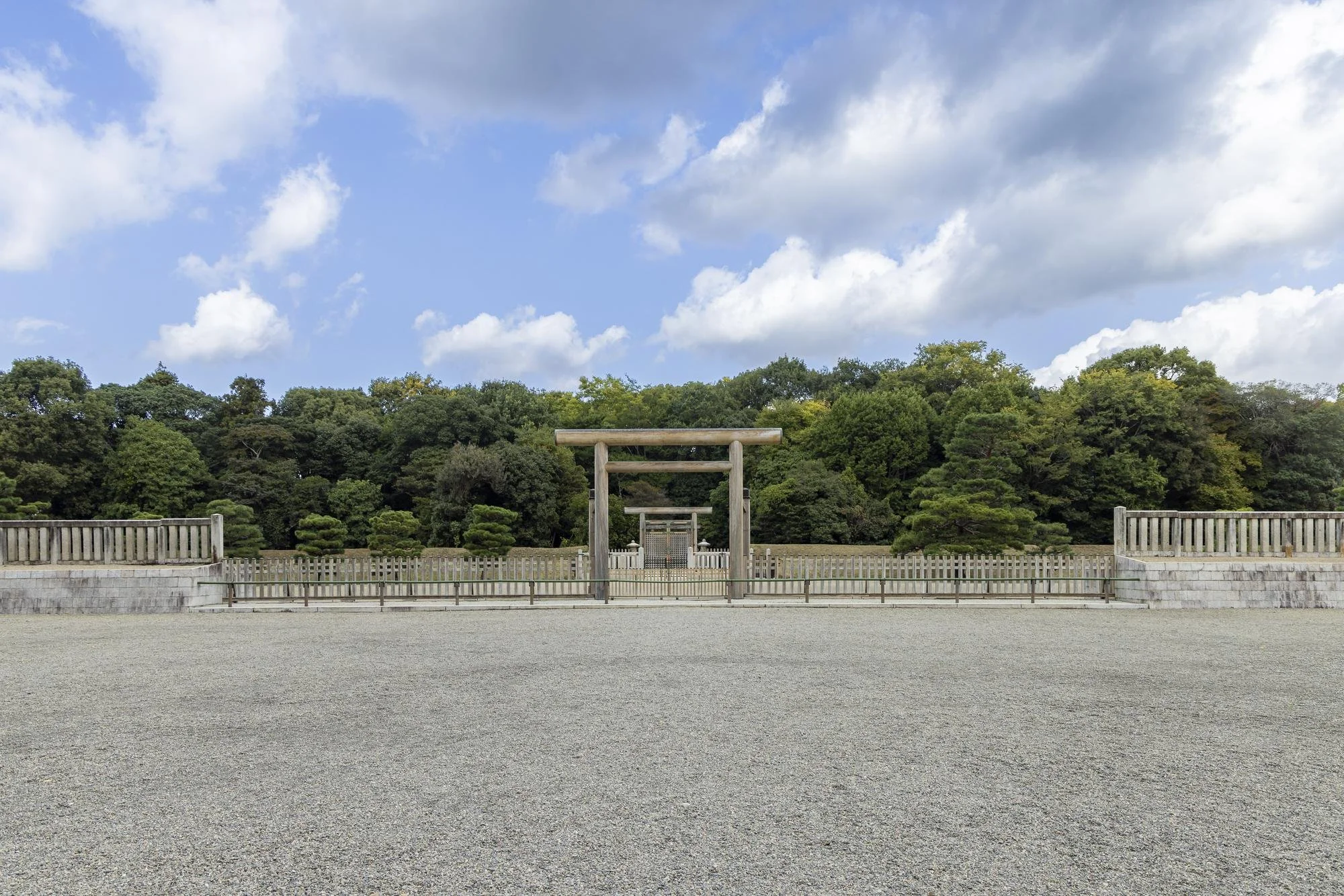 A traditional Japanese torii gate in front of a lush green garden with trees, enclosed by a wooden fence, under a partly cloudy sky.
